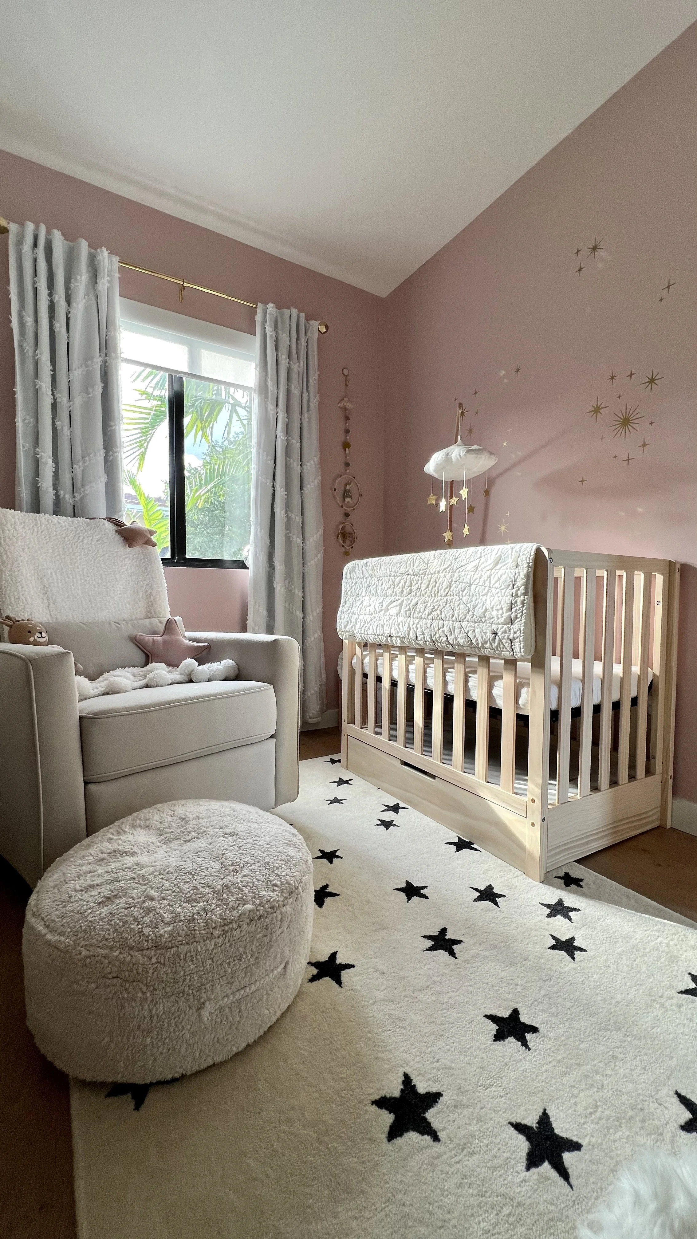 A nursery with a pink wall decorated with gold star stickers. There is a wooden crib with a white quilt, a small beige ottoman, a cream armchair with plush toys and pillows, a window with white curtains, and a beige rug with black star patterns.