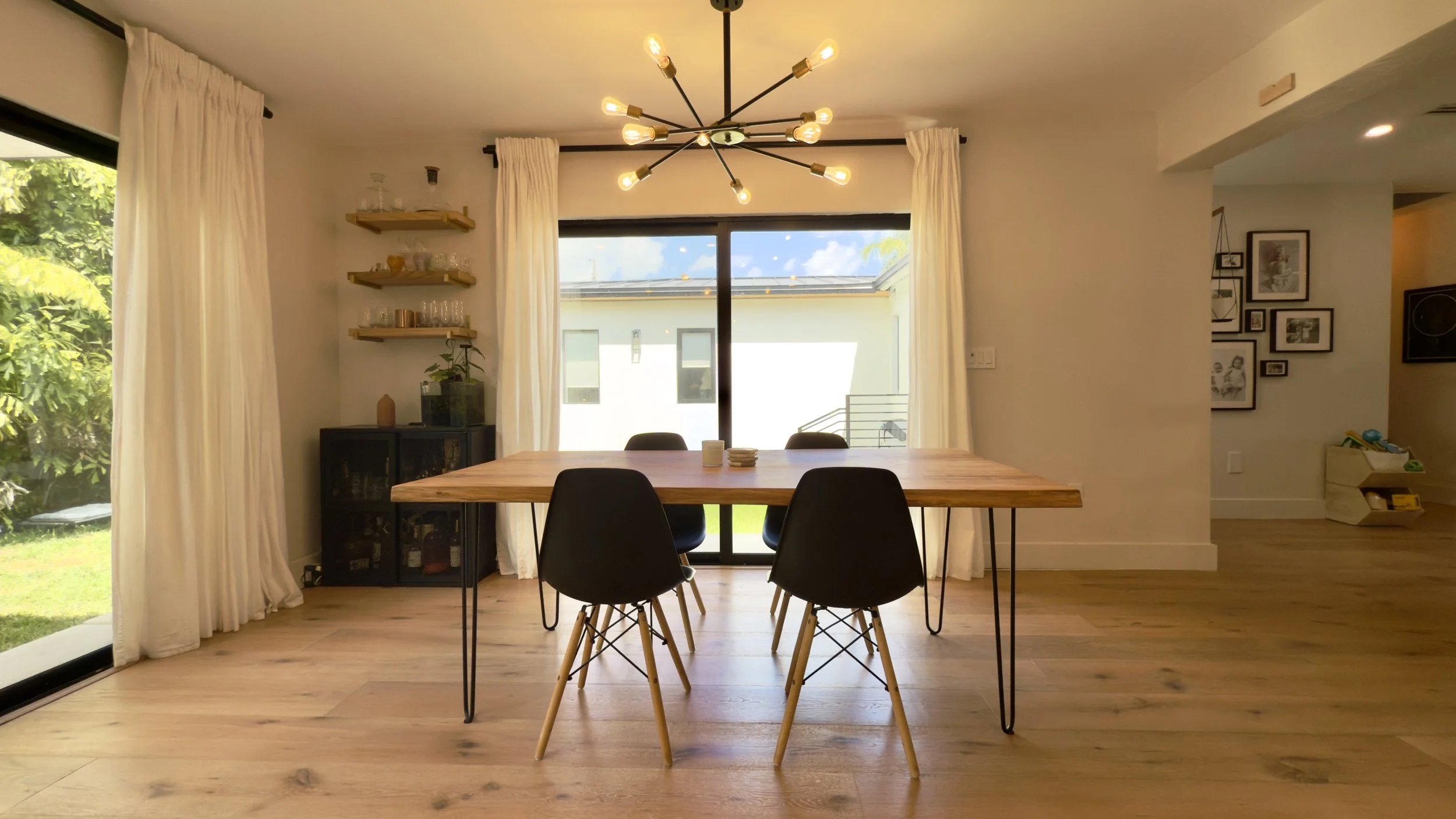 Dining room with a wooden table and four black chairs, a modern chandelier, large sliding glass doors with white curtains, and a view of a backyard with green grass and trees.