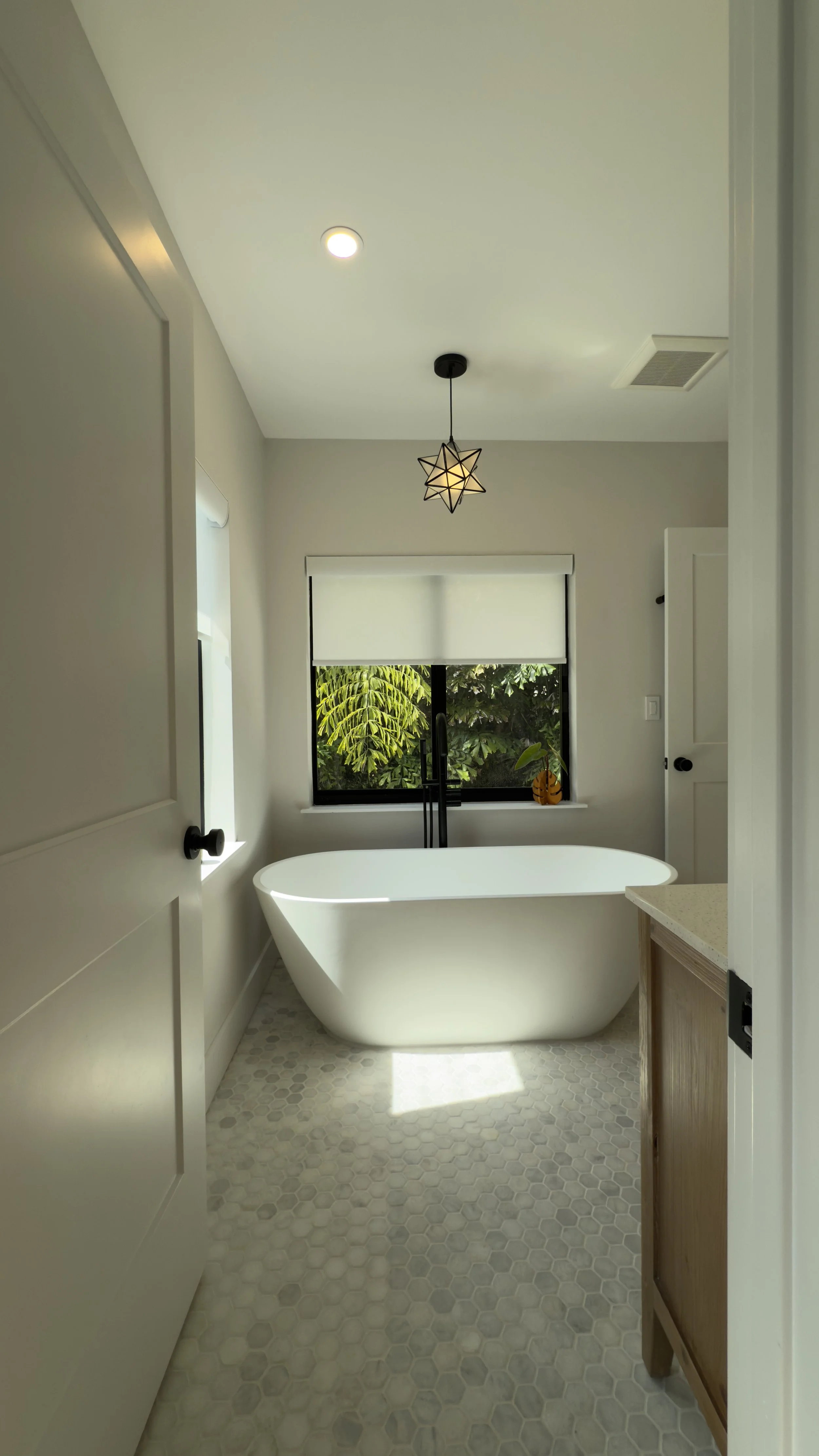 Modern bathroom with free-standing bathtub, window with greenery outside, pendant light fixture, and light-colored tile floor.