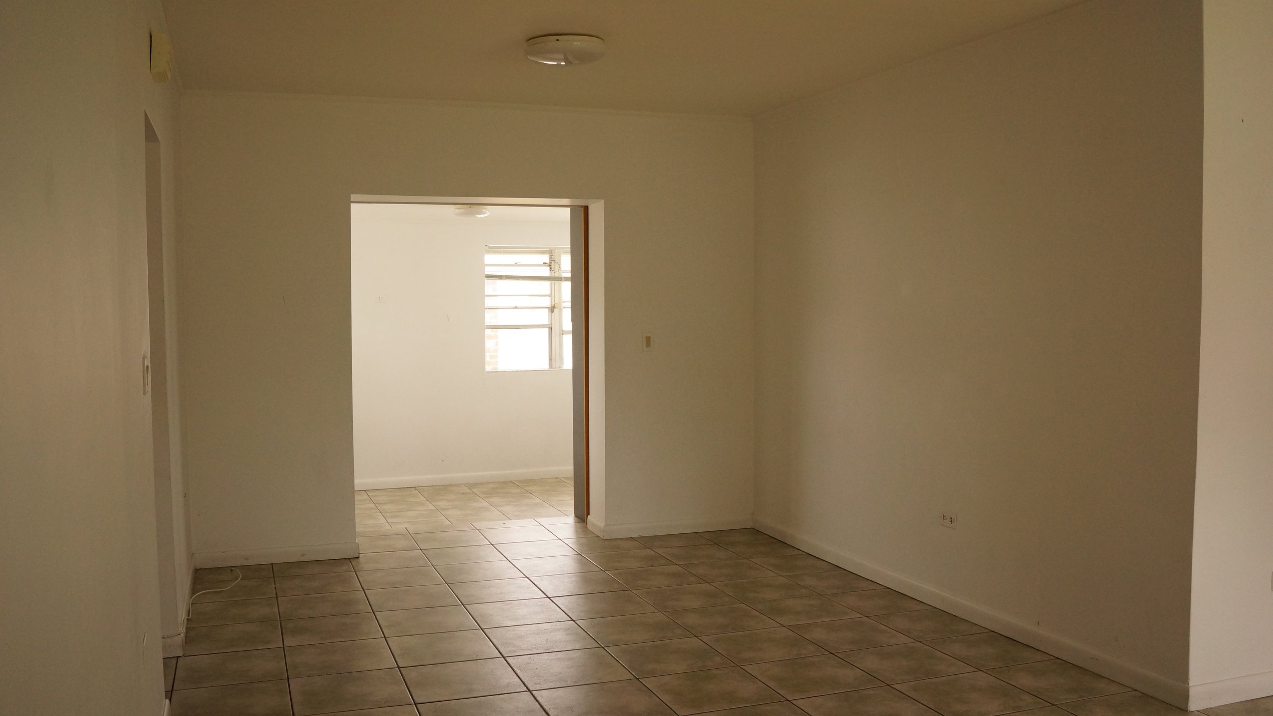 Empty room with tiled floor, white walls, a doorway to another room with a window, and ceiling lights.