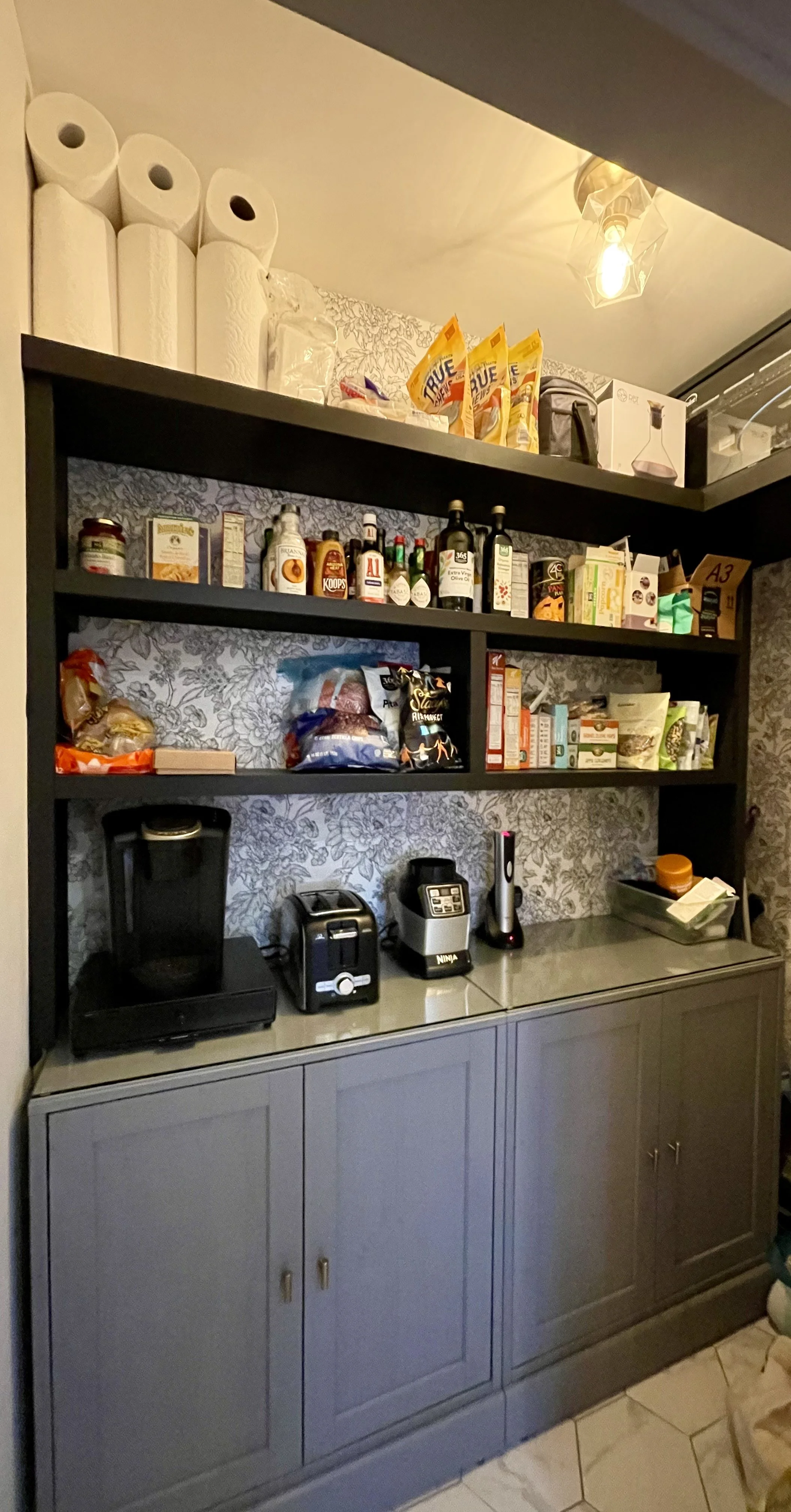 A kitchen pantry with gray cabinets and black shelves stocked with paper towels, snacks, condiments, spices, kitchen appliances, and storage containers.