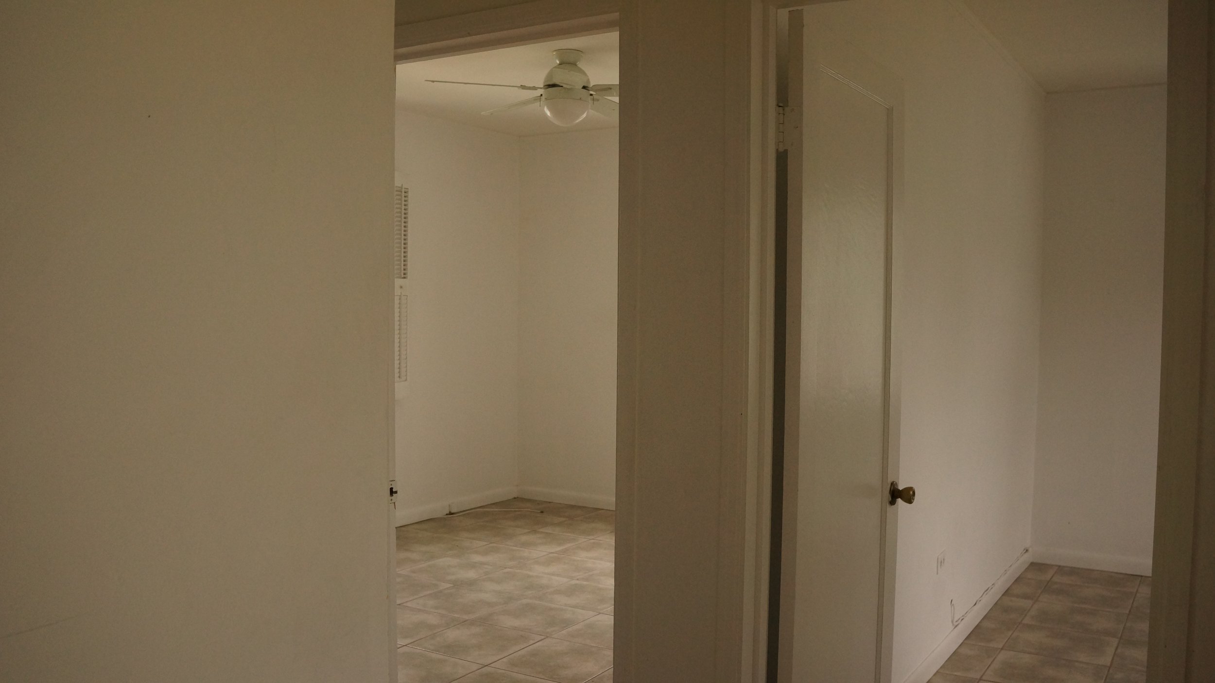 Interior view of a plain room with white walls, tiled floor, a ceiling fan with a light, and multiple doorways.