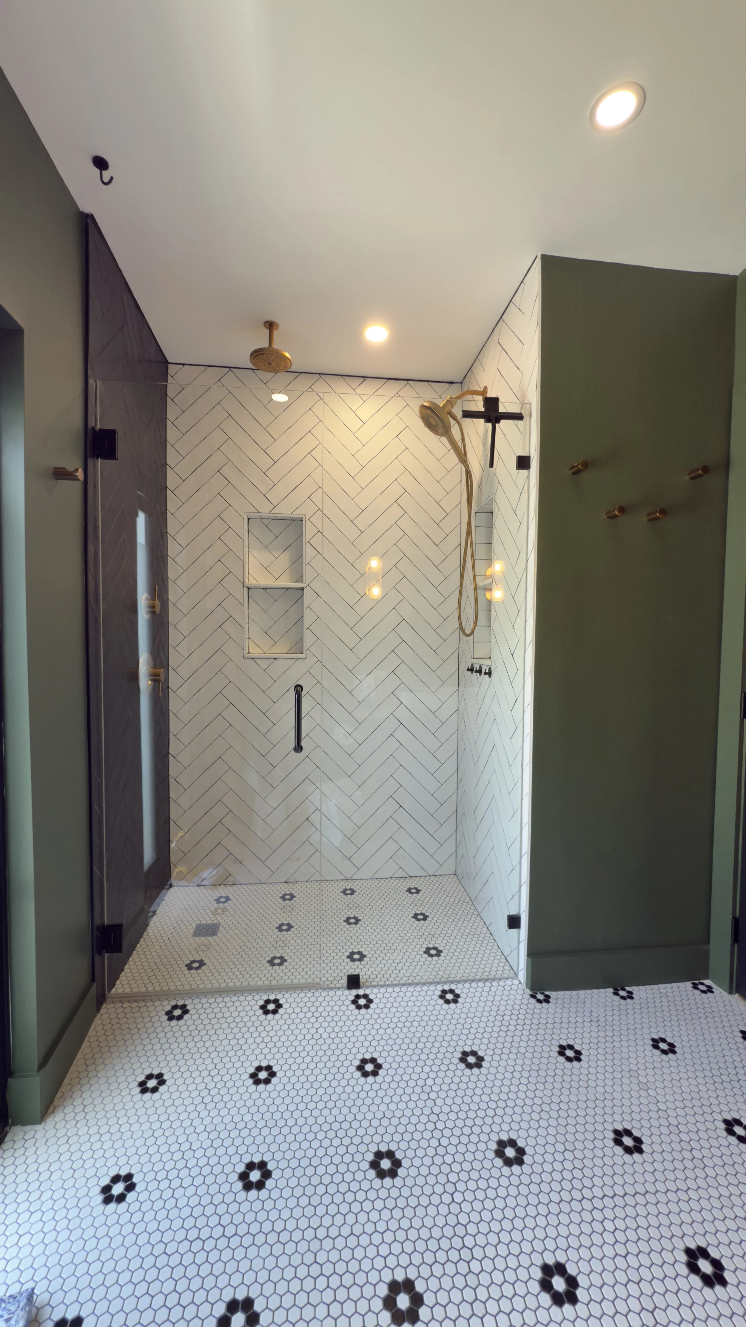 Modern walk-in shower with white herringbone tile walls, built-in shelves, a rain showerhead, and side shower fixtures, with black and white hexagon floral patterned floor tiles.