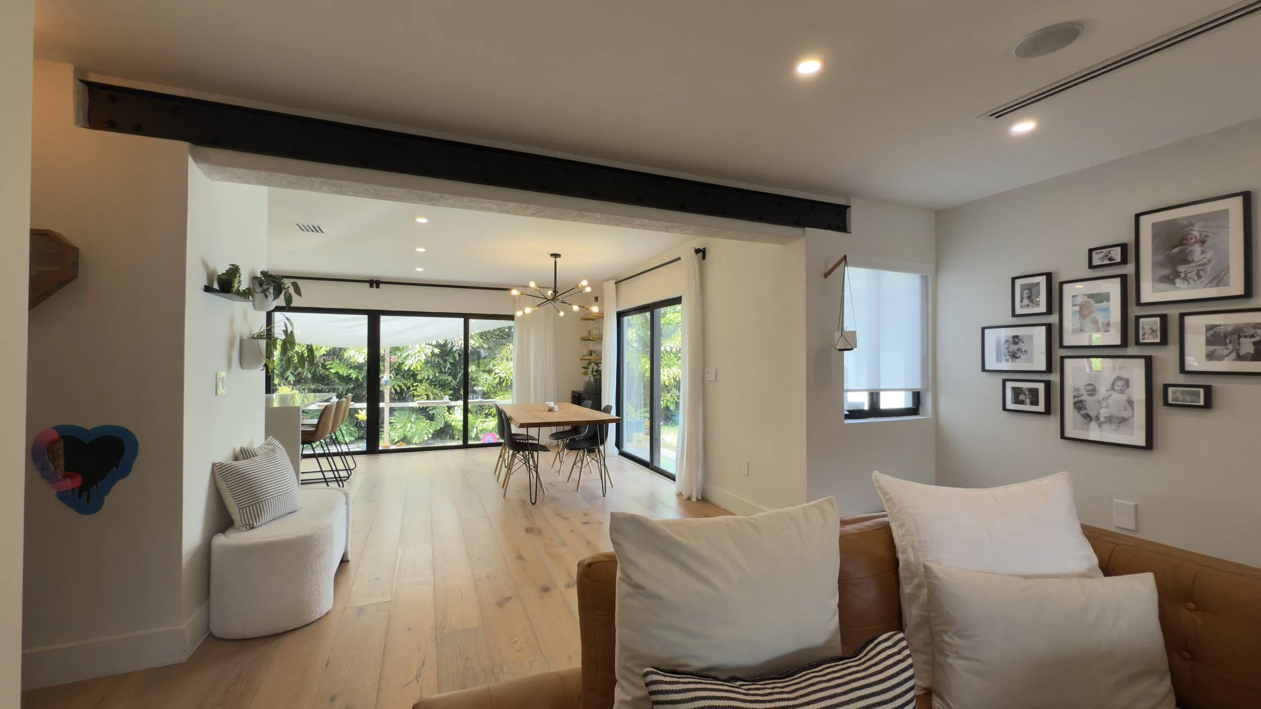 Living room with a beige couch, white pillows, wall art, and a dining area with a wooden table and black chairs, large windows showing greenery outside, and a sliding glass door leading to an outdoor balcony.