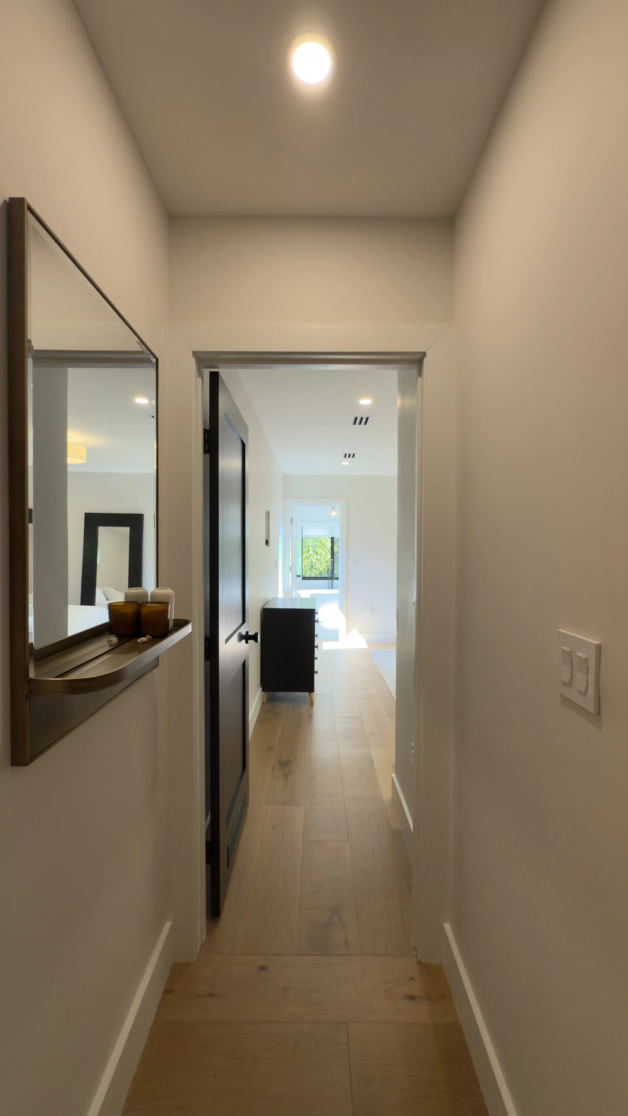 A narrow hallway leading to a living area with large windows and a balcony, natural light illuminating the space.