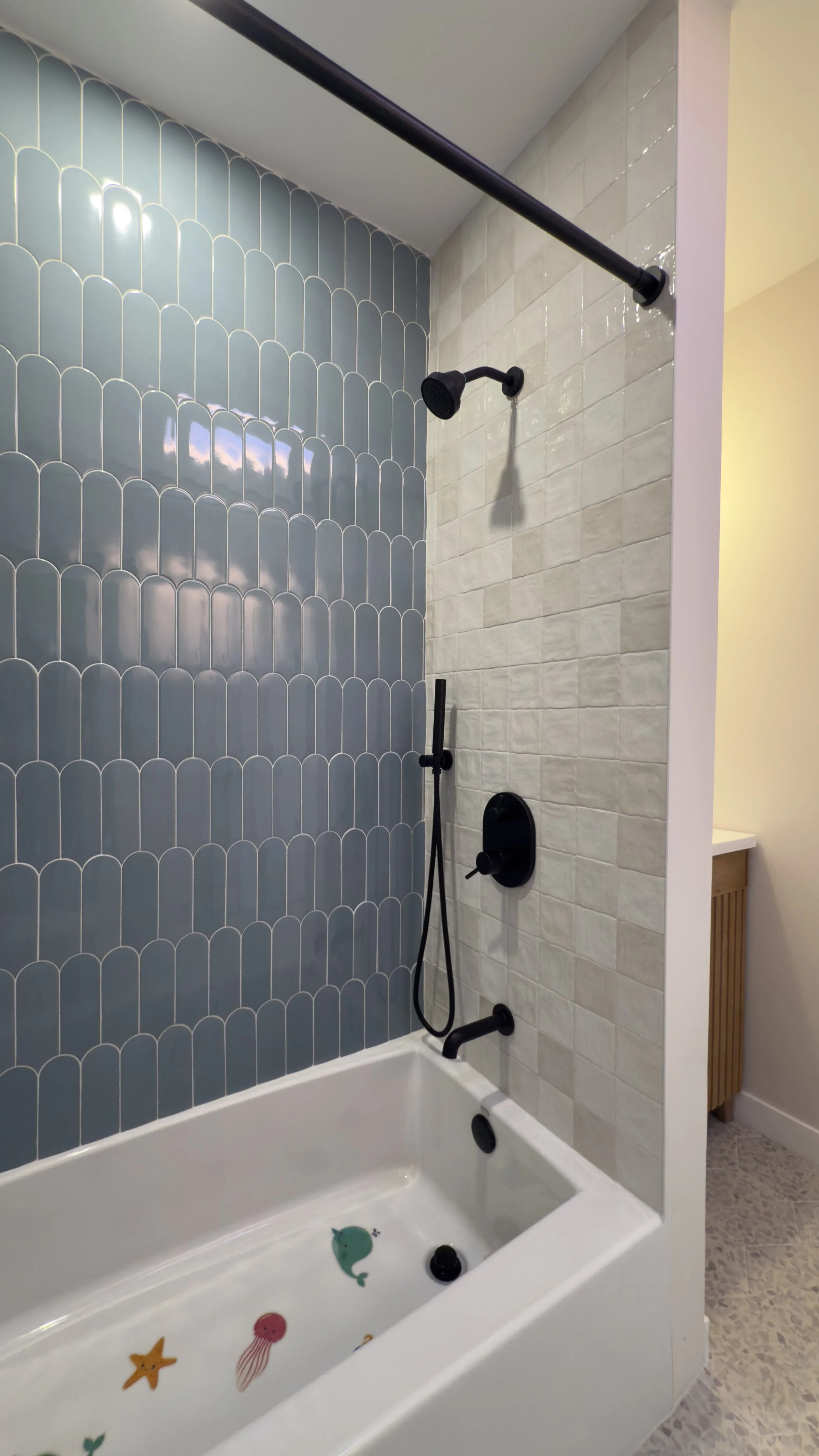 Bathroom shower with blue fish-shaped and sea creature bath toys in the bathtub, white and cream tiles on the wall, black fixtures, and a divider wall.