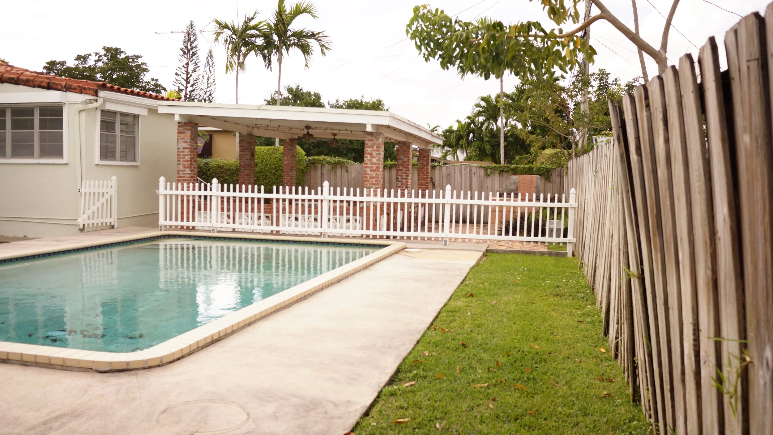 A backyard with a swimming pool, white fencing, a grassy yard, and a house with a covered patio and brick columns.