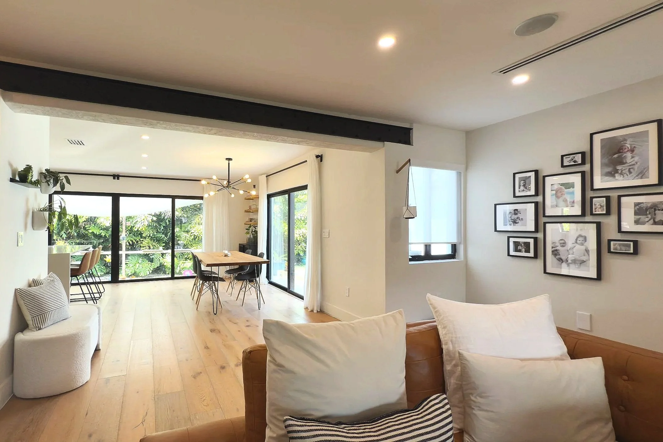 Open living and dining area with light wood flooring, white walls, a gallery wall of black-framed photos, a brown leather couch with white and striped pillows, a window with white blinds, a dining table with black chairs, large sliding glass doors leading to an outdoor garden, and modern lighting fixtures.