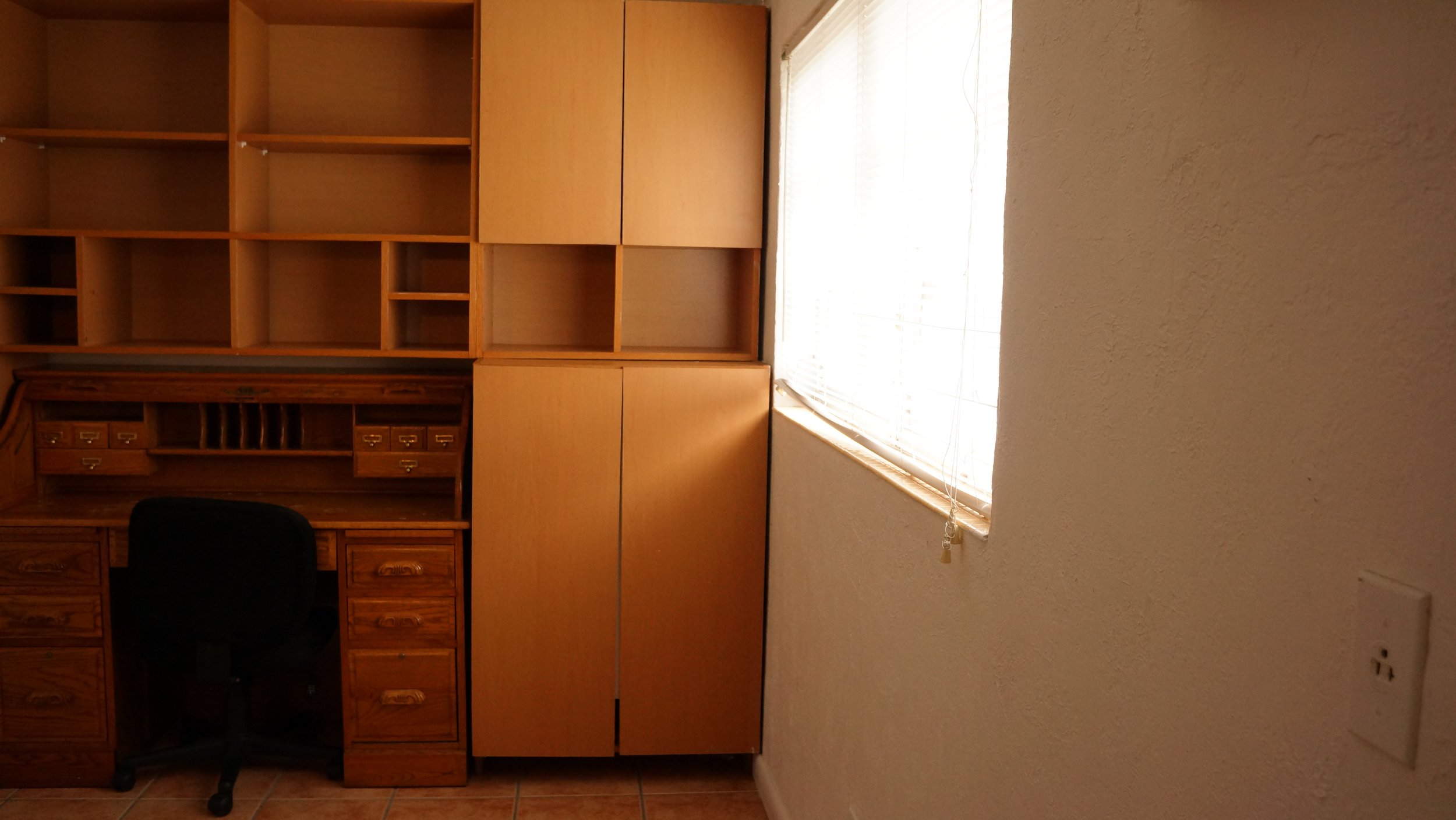 An office corner with built-in wooden desk and shelves, a black office chair, and a window with blinds allowing sunlight.