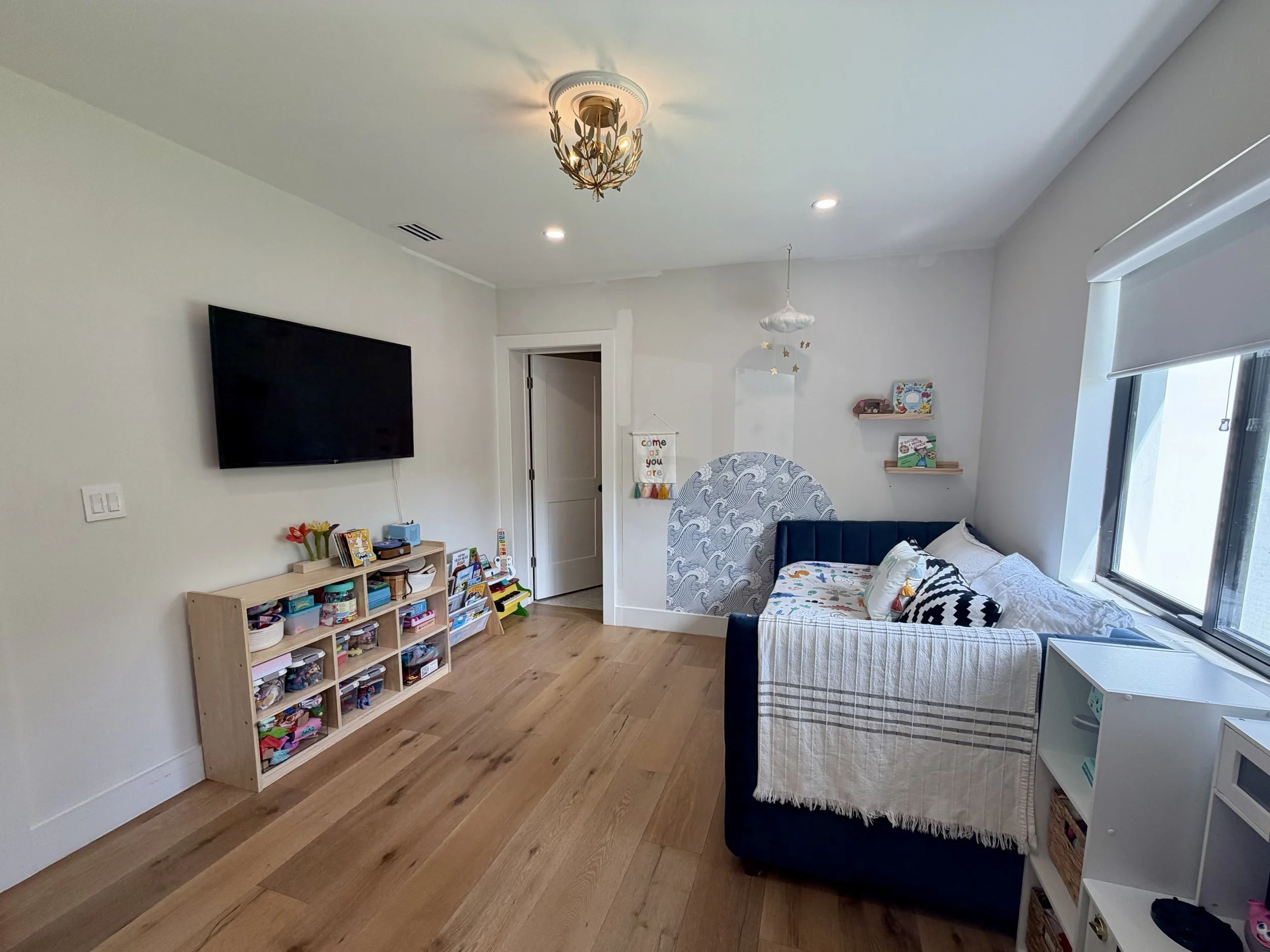 Living room with a large wall-mounted flat-screen TV, wooden shelves with toys and books, a navy sofa with white and patterned pillows, a large window, and a small white side table, with light hardwood floors and a decorative ceiling light.
