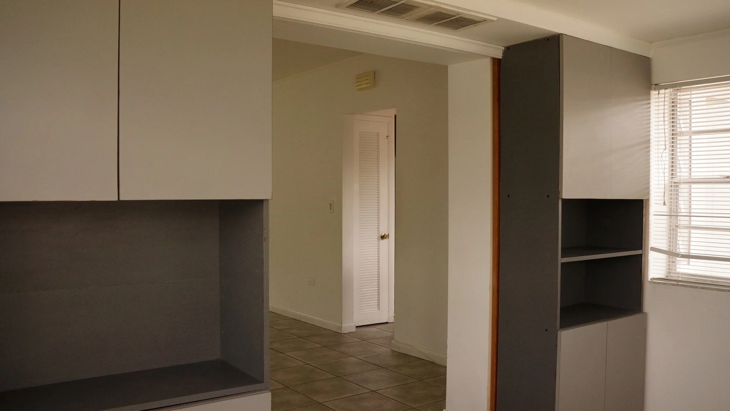 Interior view of a room with built-in cabinets, tiled floor, a door with a louvered panel, and a window with blinds.