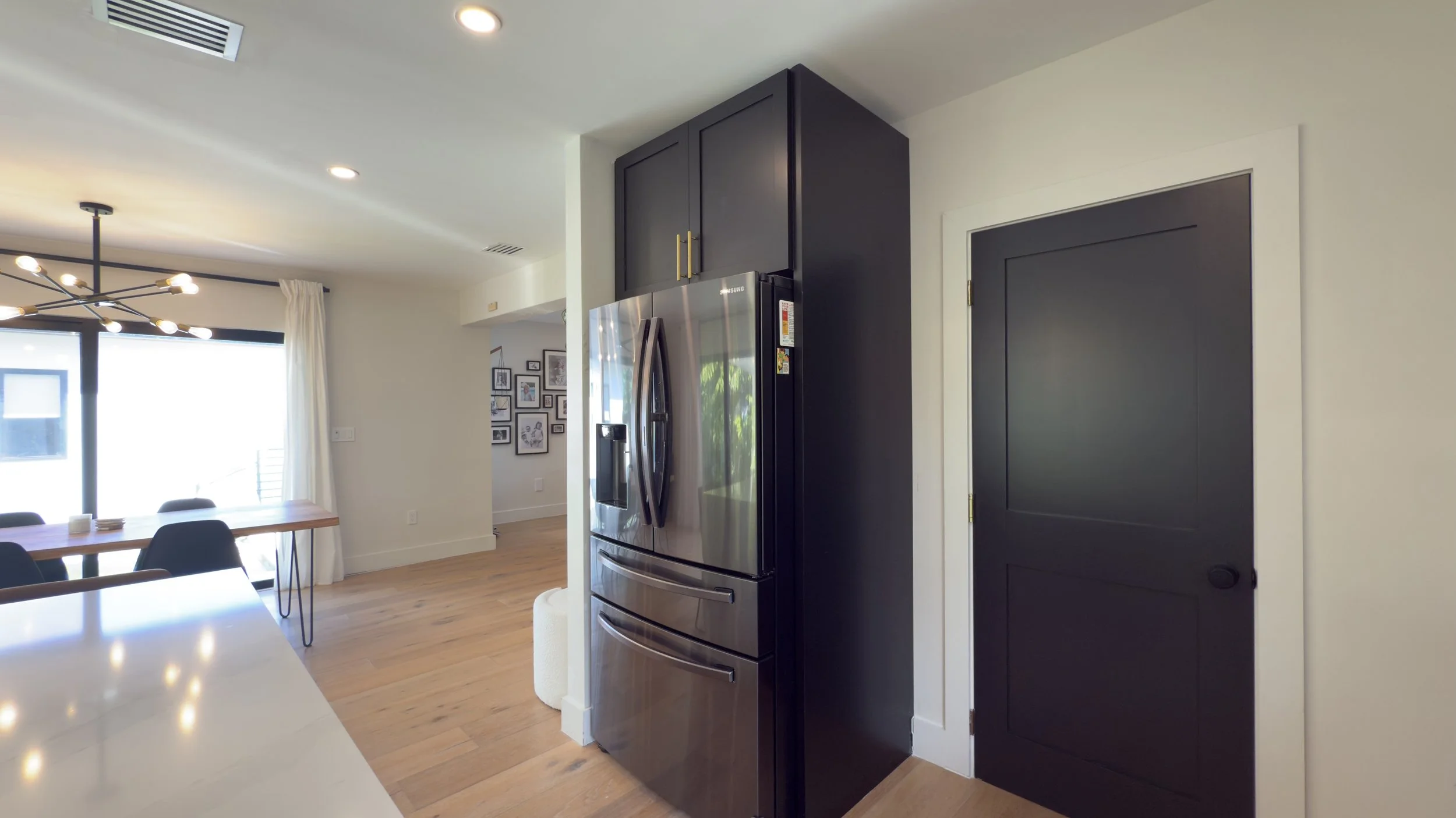 Modern kitchen with stainless steel refrigerator, black cabinets, and a black door, with hardwood flooring and a dining area with black chairs and large window.