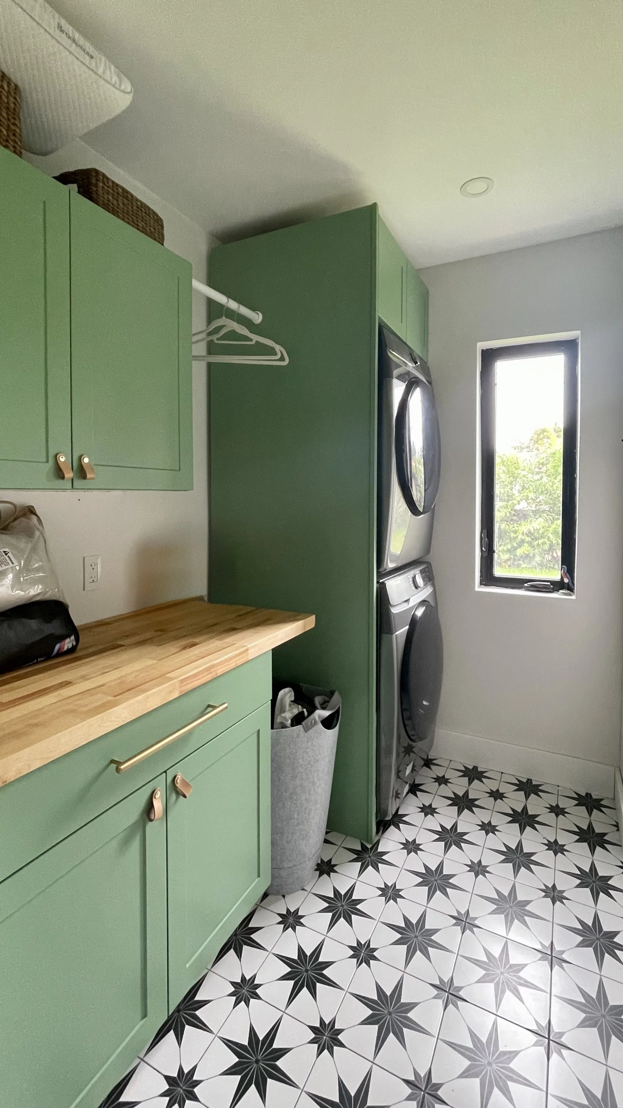 Laundry room with green cabinets, a stacked washer and dryer, a window, black and white star-patterned floor, a trash bin, and a wooden countertop.