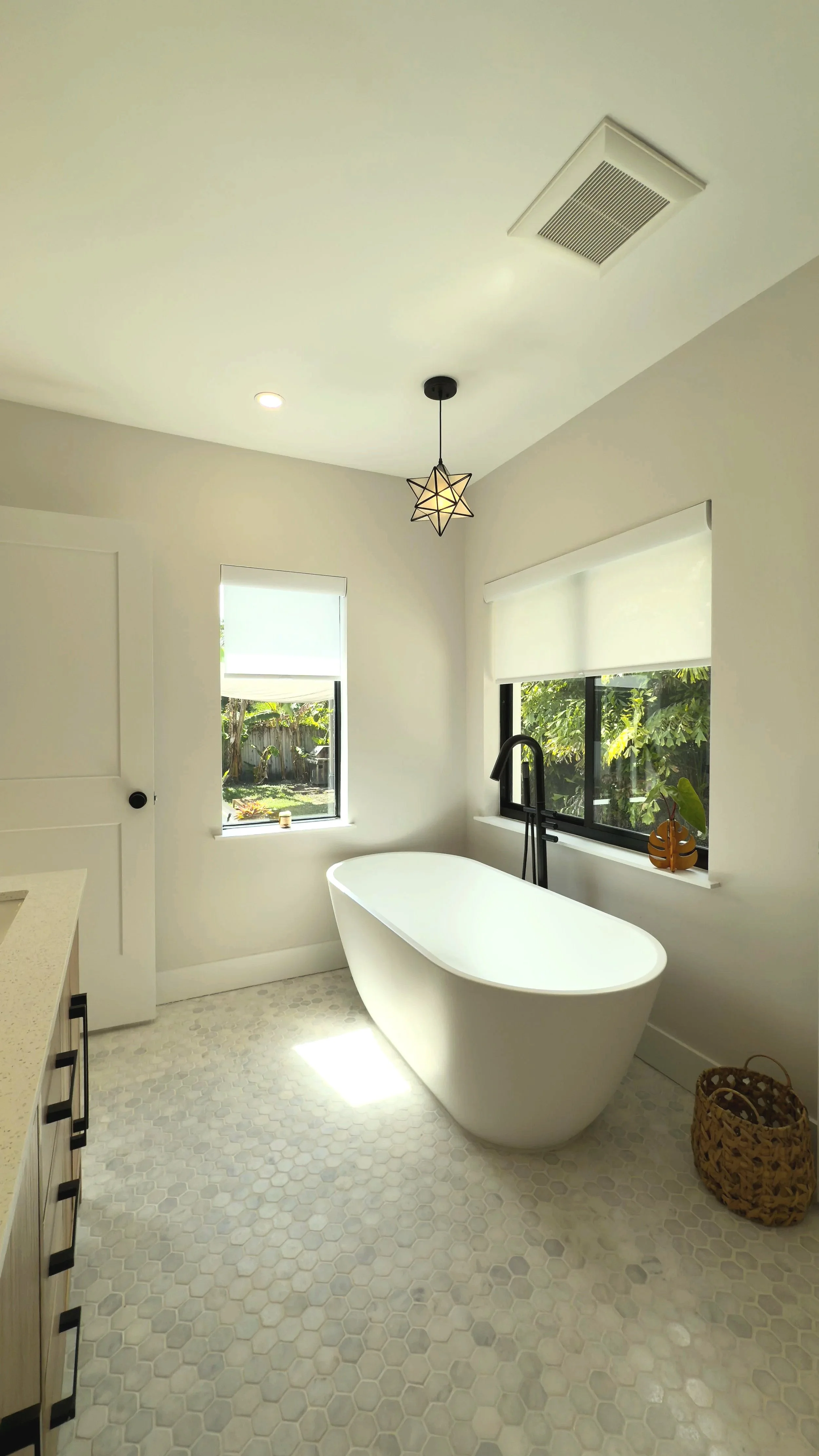 Modern bathroom with a freestanding bathtub, two windows with blinds, a black floor faucet, a wicker basket, and a decorative pendant light