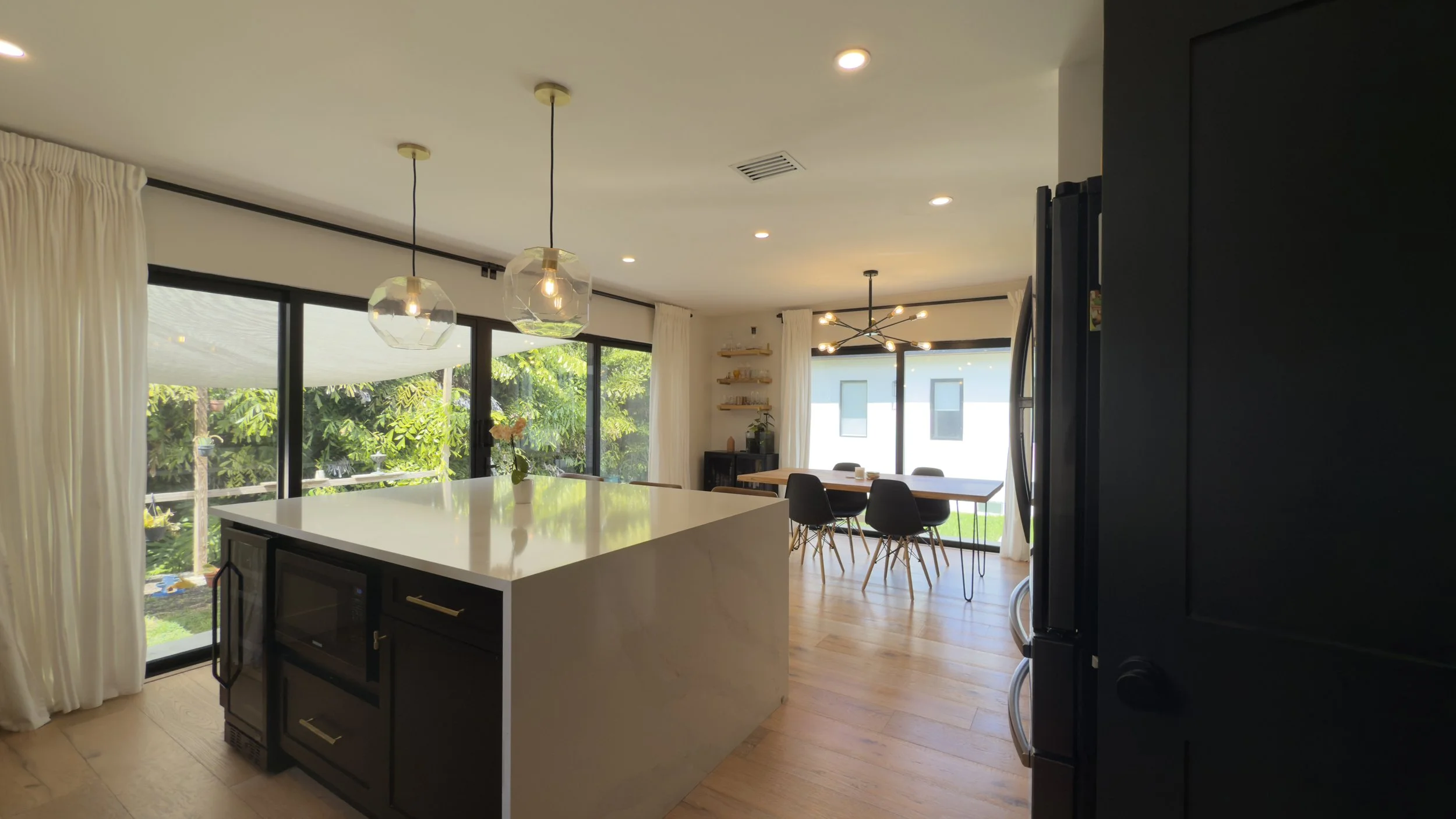 Modern open-concept kitchen and dining area with large windows, white walls, wooden floors, a kitchen island with a white countertop, black cabinetry, pendant lights, a dining table with black chairs, and outdoor greenery visible through the windows.