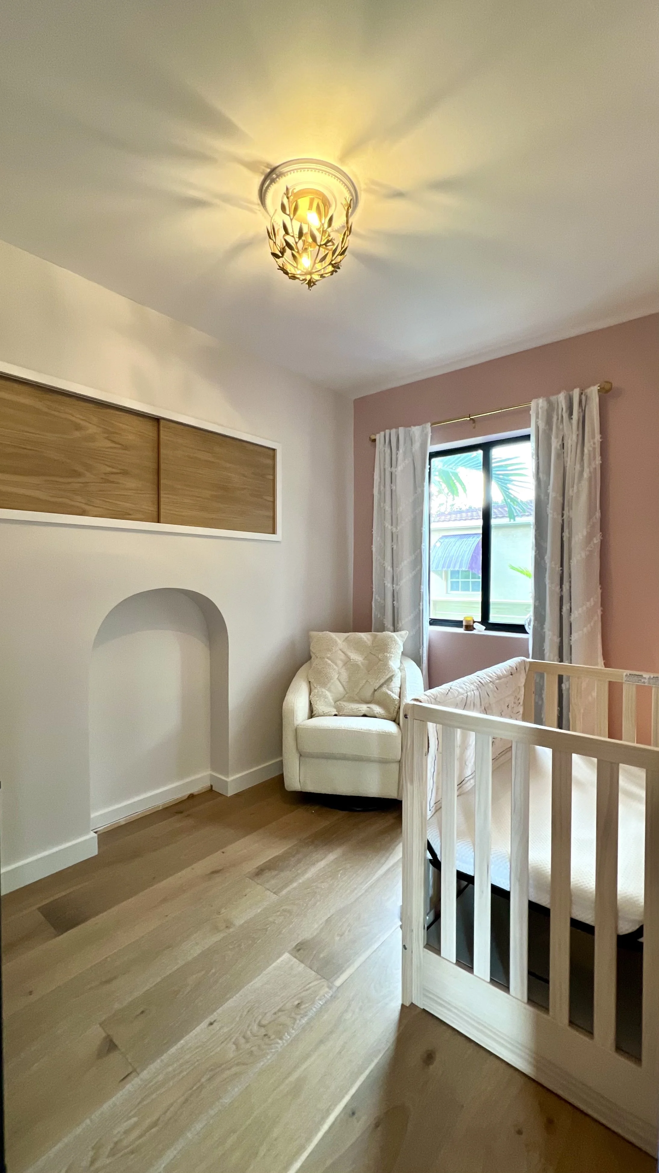 A cozy nursery room with wooden flooring, a white armchair, a crib, a window with white curtains, a pink accent wall, and a ceiling light fixture.