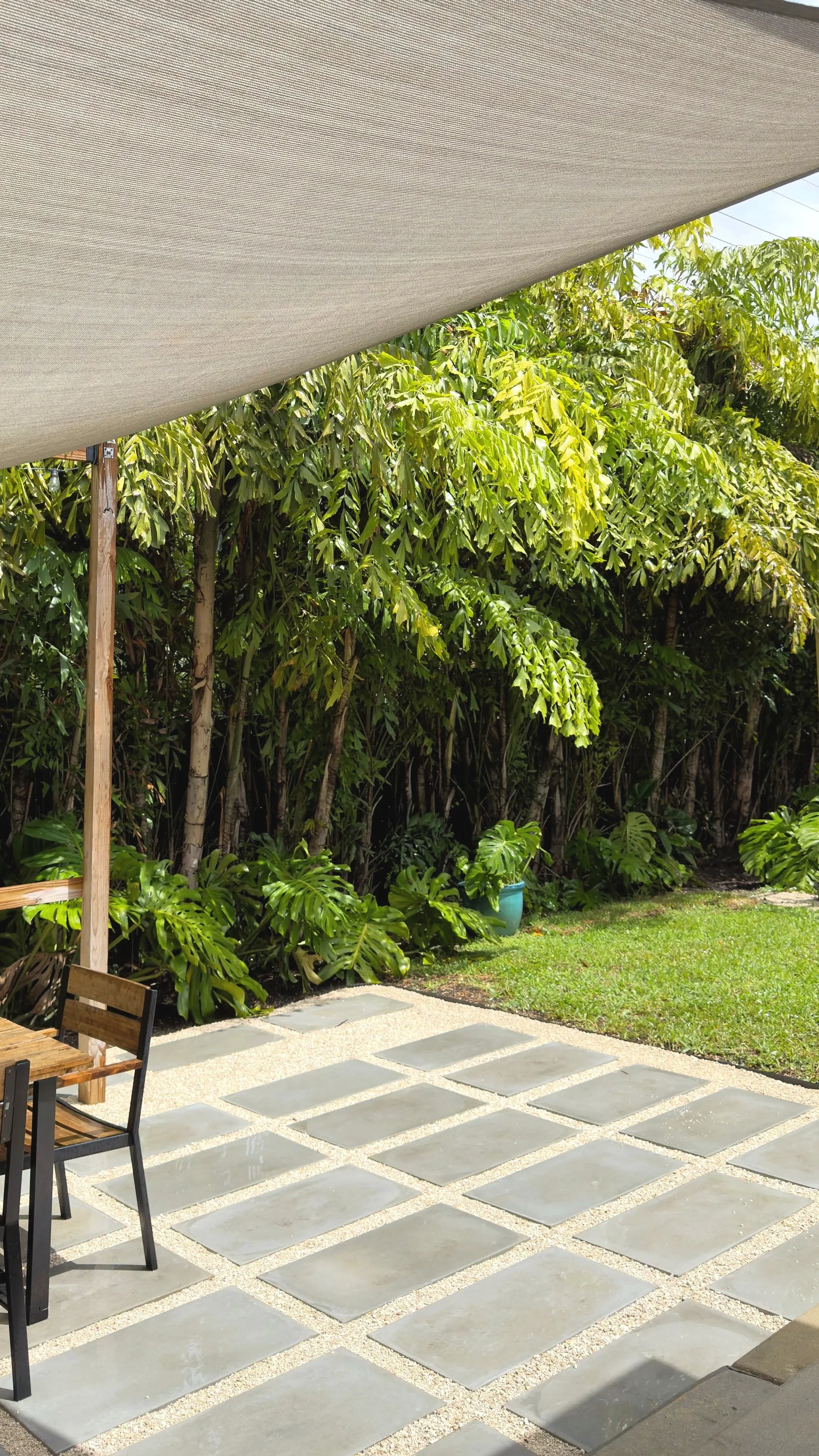Outdoor patio area with stone pavers, partially shaded by a large fabric canopy, surrounded by lush green tropical plants.