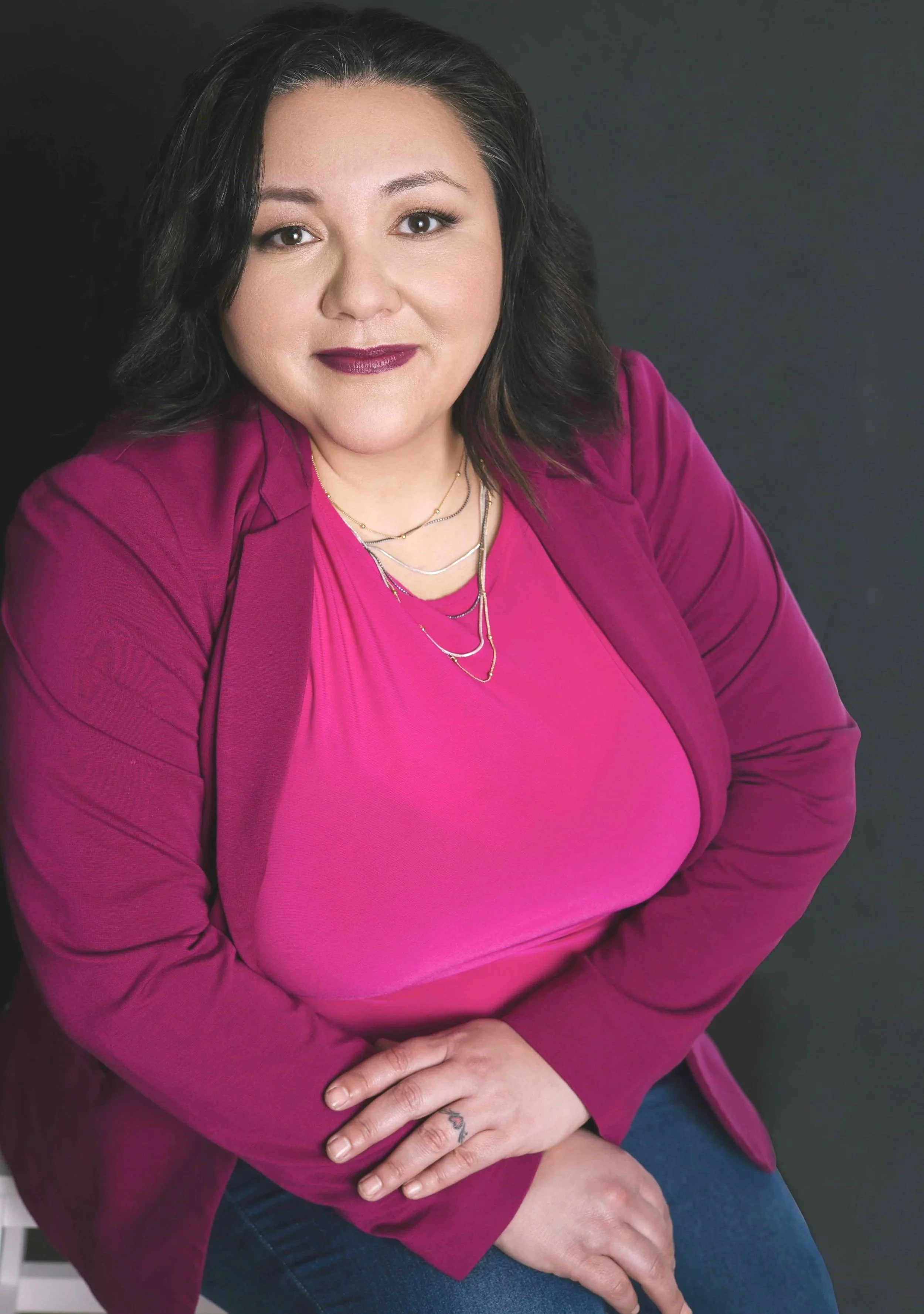 A woman with dark hair and light skin sitting against a dark background, wearing a magenta blazer and matching top, with layered necklaces, with her arms crossed.