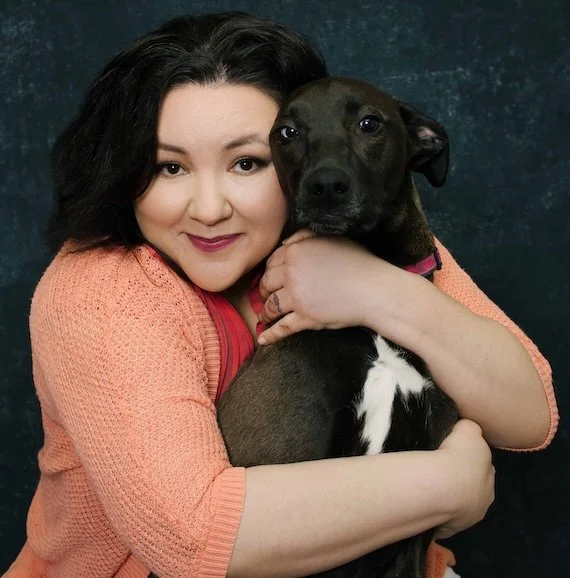 A woman with black hair and a peach-colored sweater hugging a brown and black dog with a white patch on its chest, against a dark background.