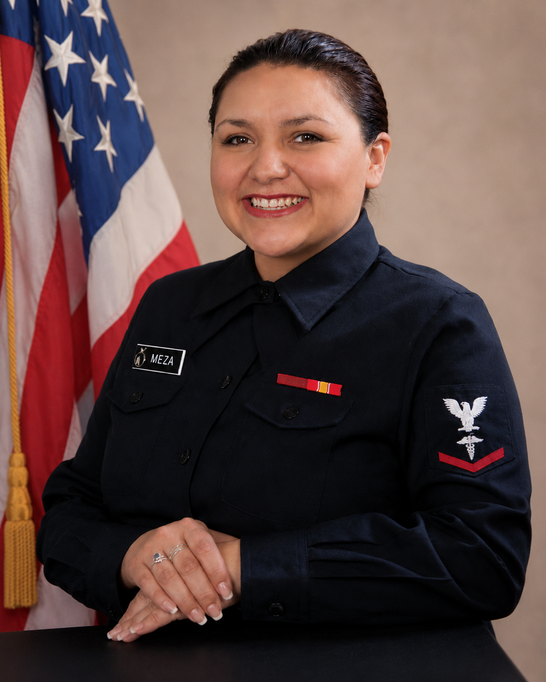 A female police officer in uniform with a name tag reading 'Meza,' standing in front of an American flag, smiling with hands clasped.
