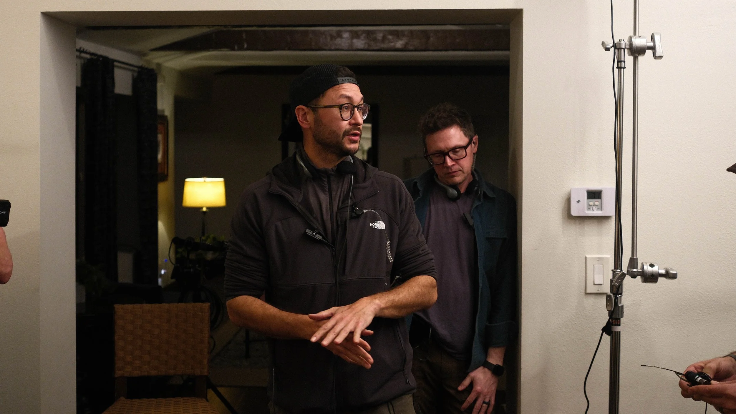 Two men standing in a room, engaged in conversation or discussion, with equipment and a lamp in the background.