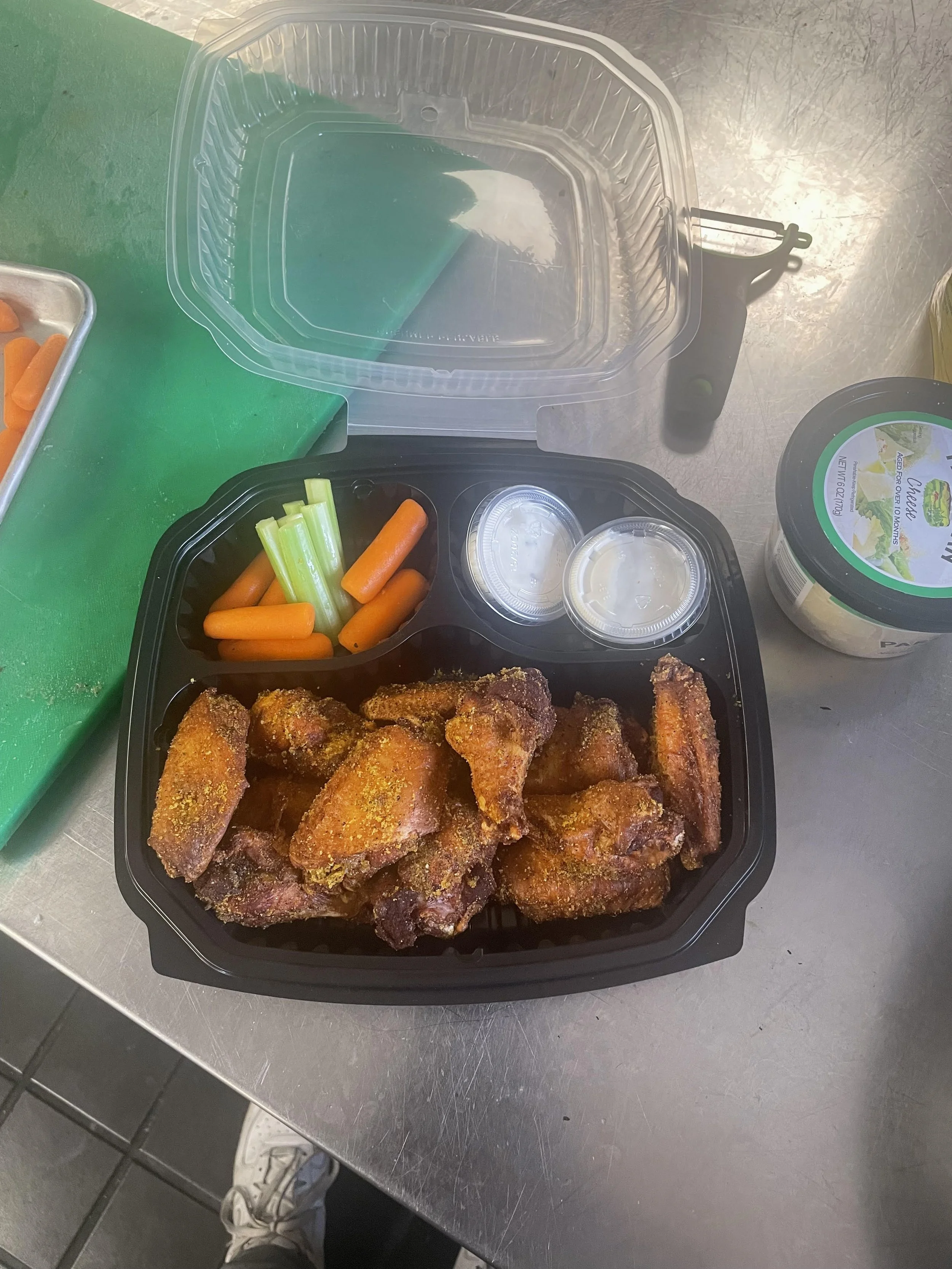 A black plastic takeout container with fried chicken wings, celery sticks, and baby carrots, paired with two small containers of dipping sauce. There's a jar of mayonnaise-type spread and a green cutting board with additional pieces of carrots in the background.