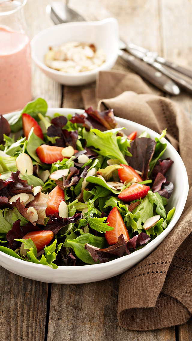 Fresh mixed greens salad with strawberries and sliced almonds in a white bowl on a wooden table.