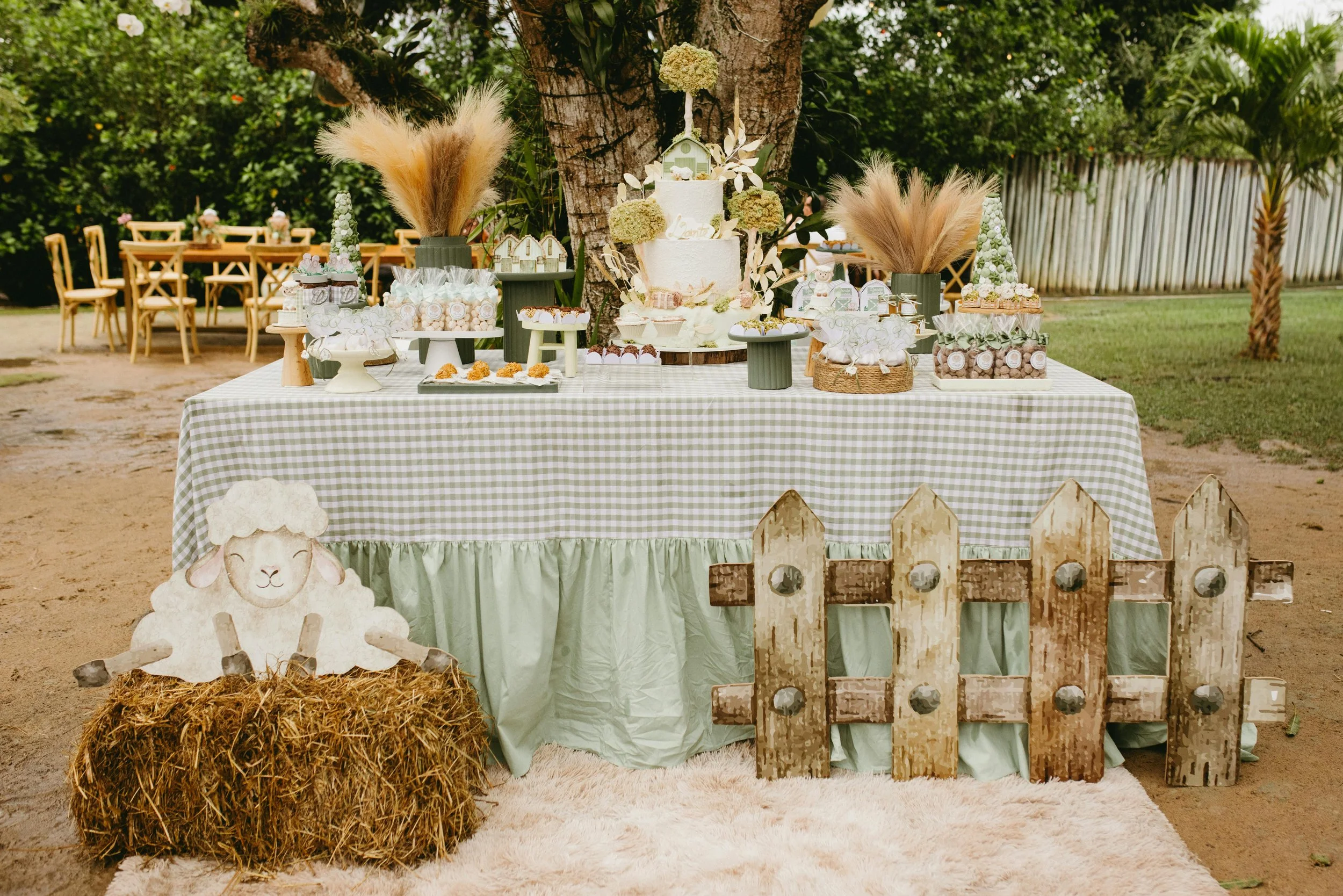 A decorated outdoor table with a green and white checkered tablecloth, featuring a two-tiered white cake, cupcakes, and various sweets, set under a large tree. The area has rustic wood and hay accents, surrounded by chairs and greenery.