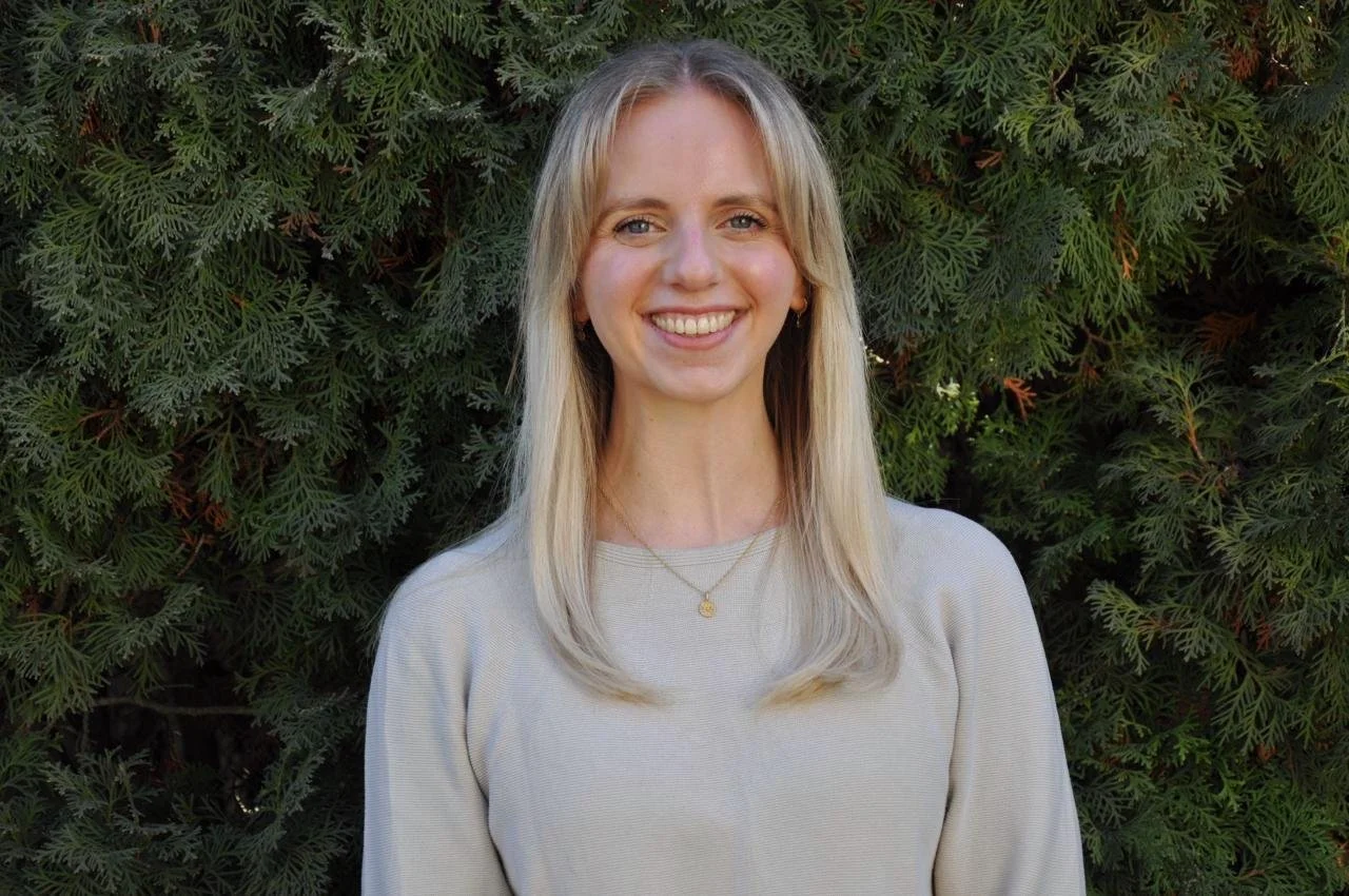 A young woman with long blonde hair, smiling, standing outdoors in front of green leafy bushes.