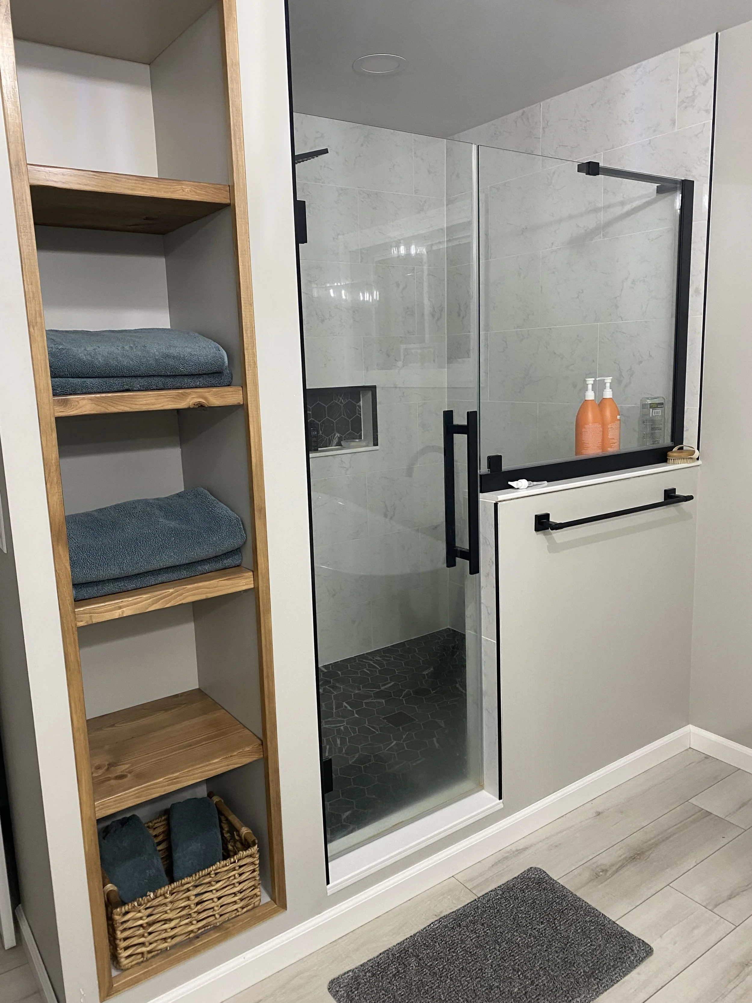A bathroom with a glass shower enclosure, a wooden shelving unit with towels and a basket, and a gray bath mat on light-colored wood flooring.