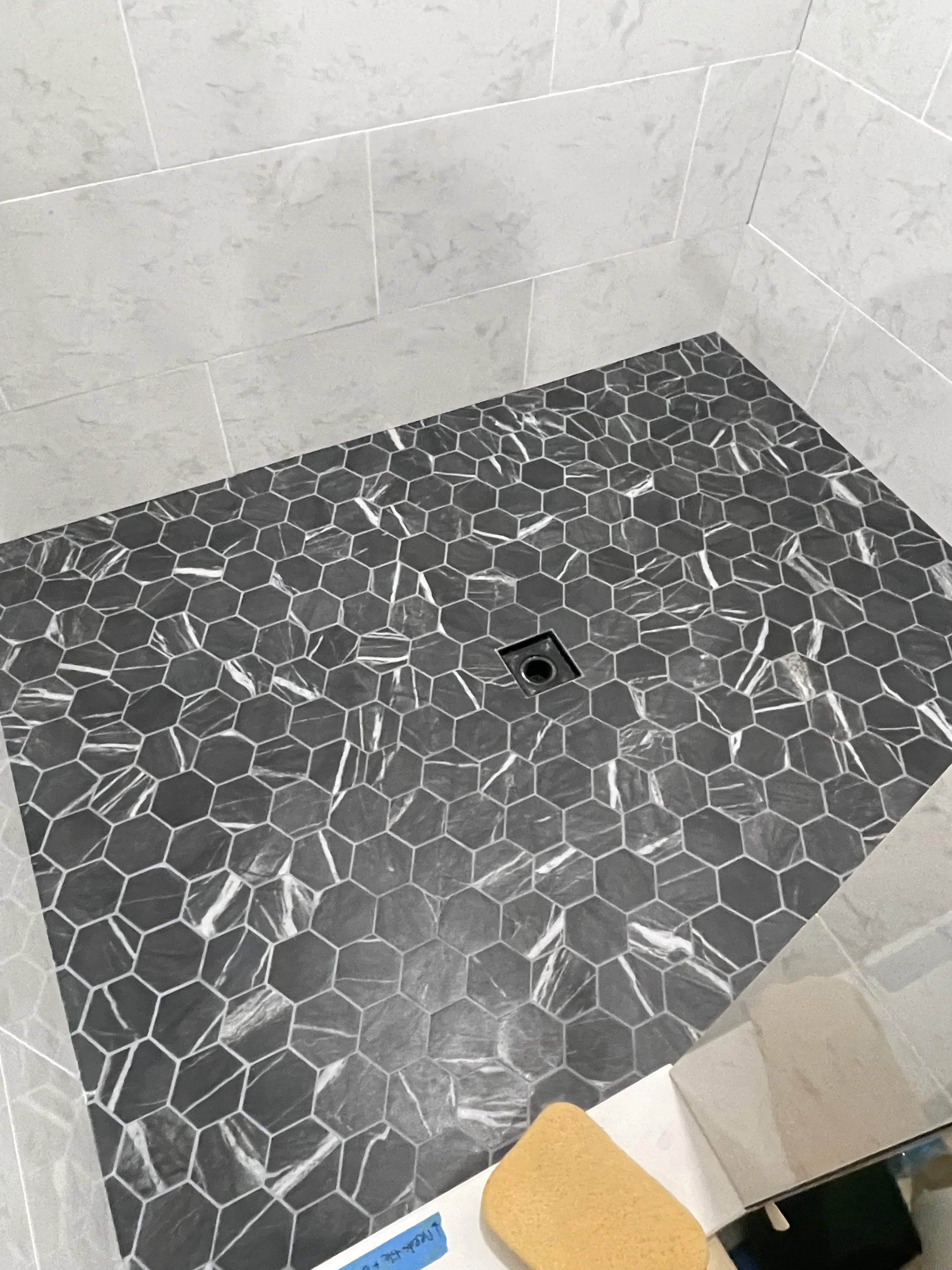 Tile shower floor with hexagon black and white marble pattern, wall with light gray tiles, drain in the center, with a yellow sponge at the edge.