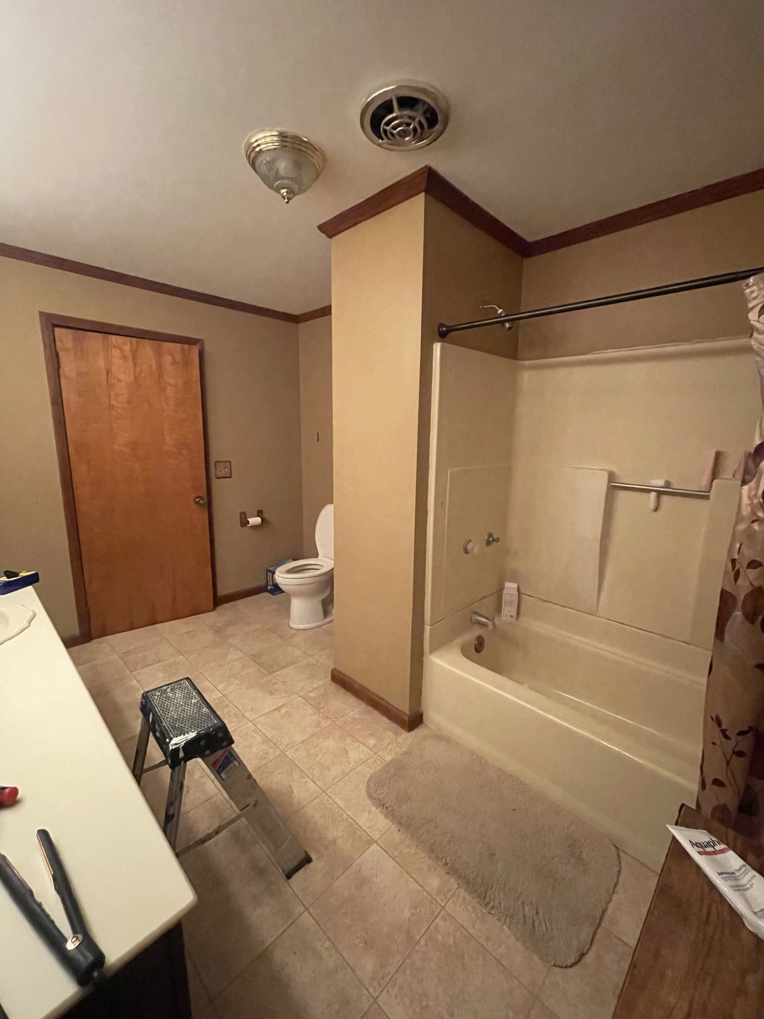 A bathroom with a toilet, bathtub, and shower, with beige tiled floor and tan walls, wood door, ceiling light, and a small stool.