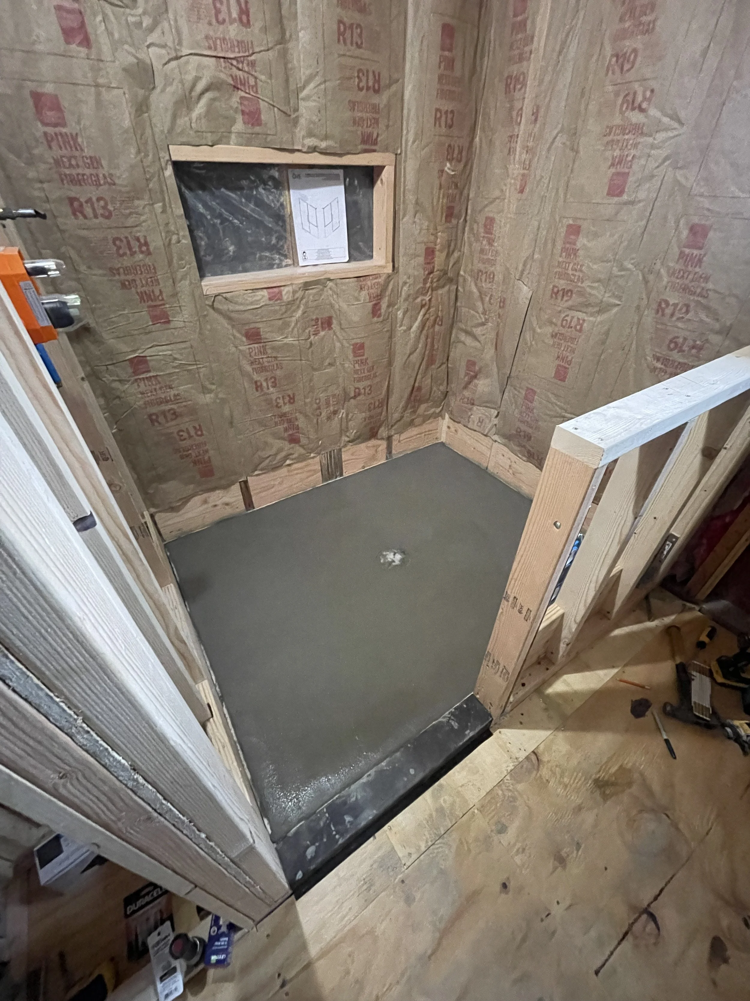 A small bathroom under construction with a concrete floor and unfinished walls covered with insulation. There is a small window on the wall and a punch in the concrete floor. Construction tools and materials are visible nearby.