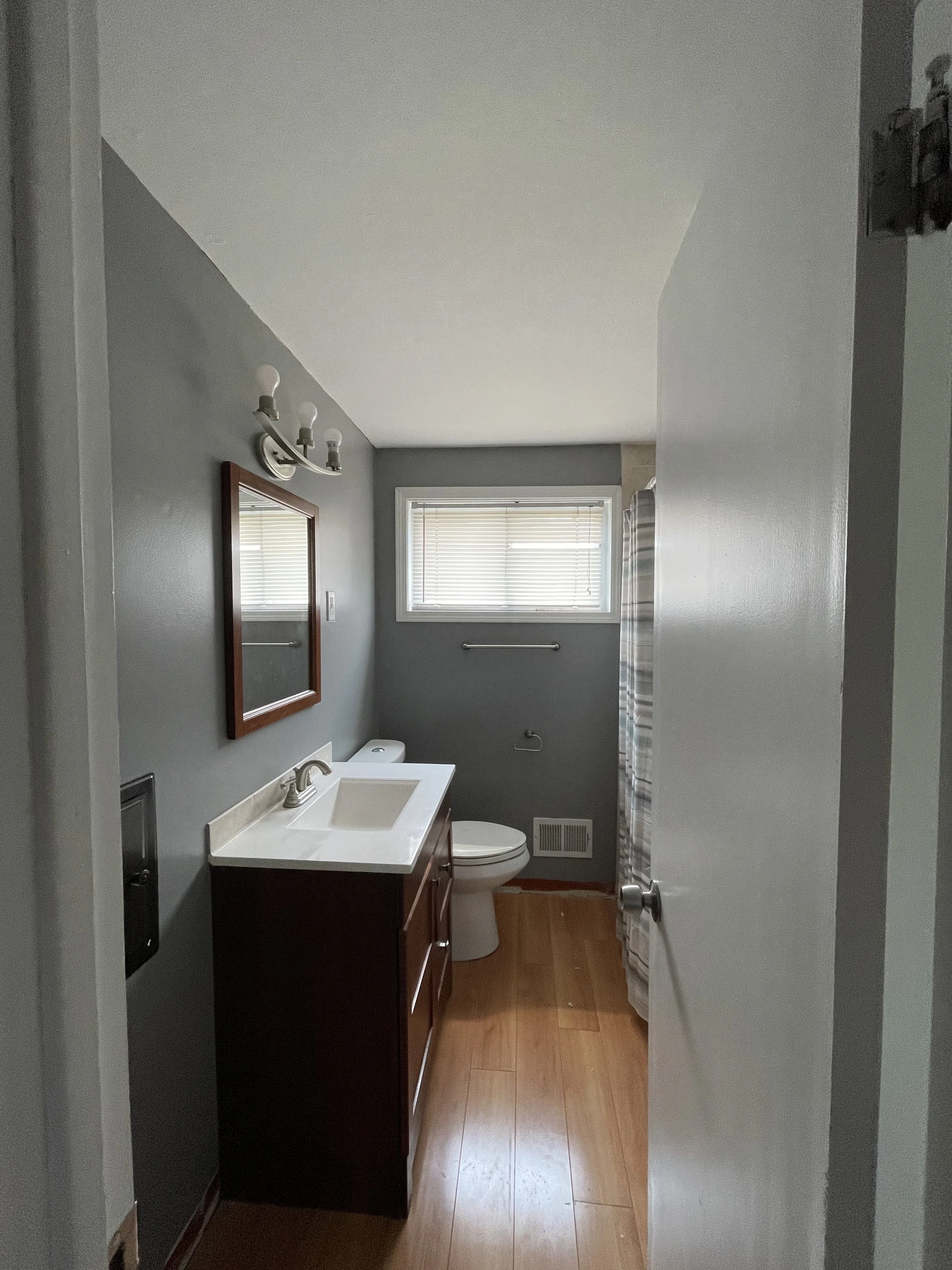 A small bathroom with gray walls and hardwood floors, featuring a wooden vanity with a white sink, a mirror above, a wall-mounted light fixture, a toilet, a window with blinds, and a shower with a striped curtain.