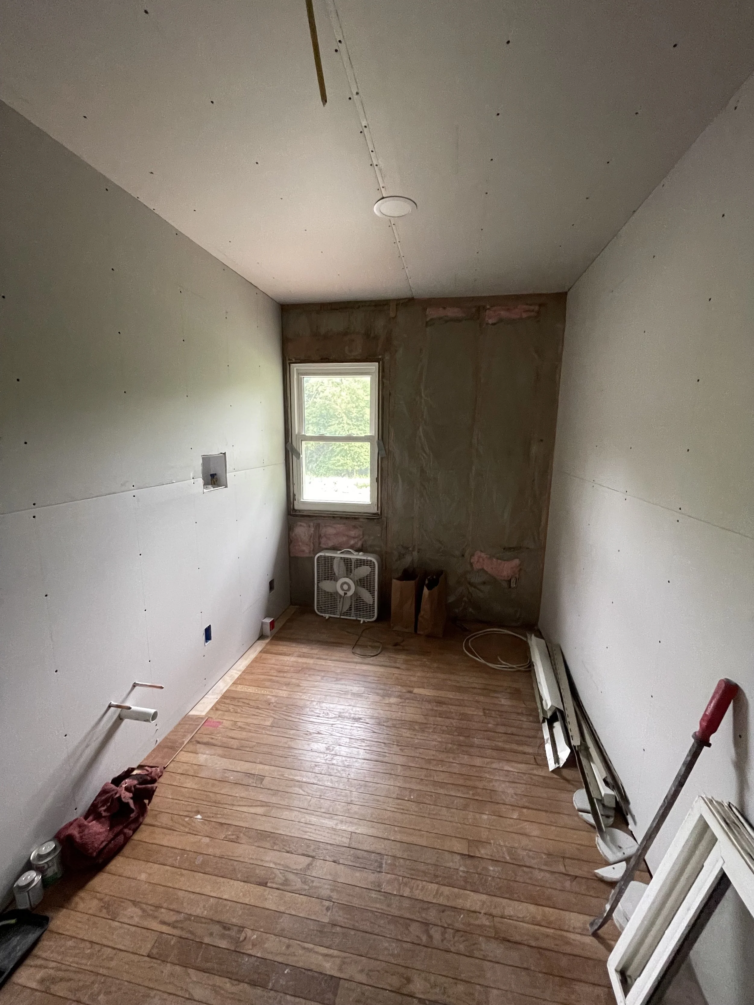 A small room under renovation with hardwood floors, a window, and unfinished walls. There are construction tools and materials on the floor, including a fan, wood trim, and a ladder.