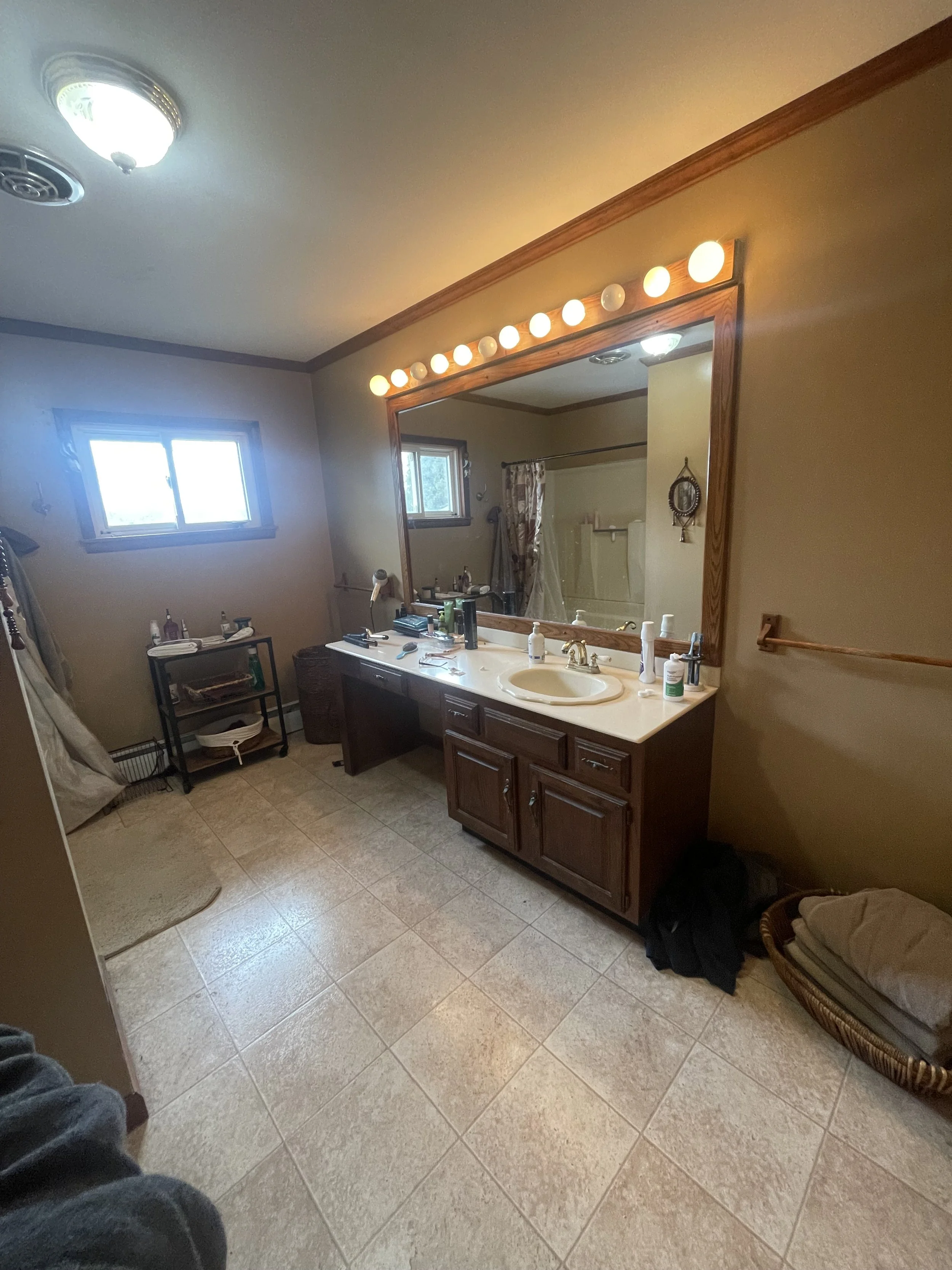 Bathroom with a large mirror above a double sink vanity, vanity is cluttered with toiletries, small window, ceiling light, and tiled floor.