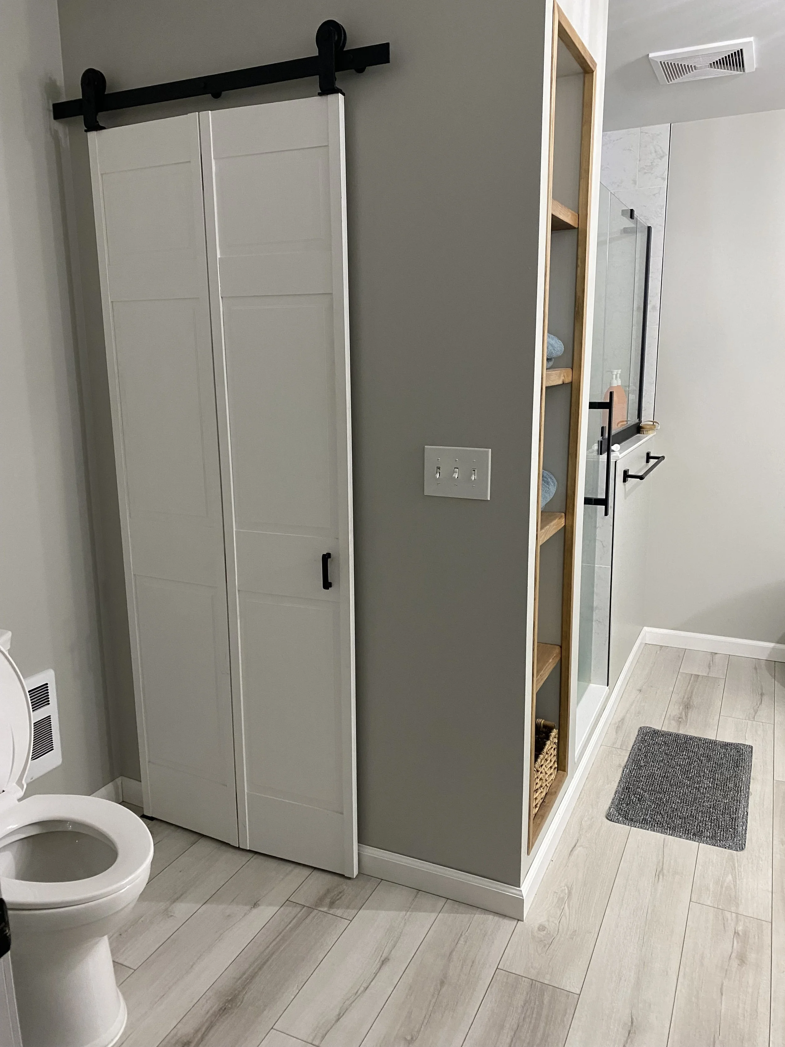 Bathroom with a white toilet, a sliding closet door, a gray wall, a wooden shelving unit, a shower area with a glass door, a towel bar, and a gray rug on the light-colored wood floor.