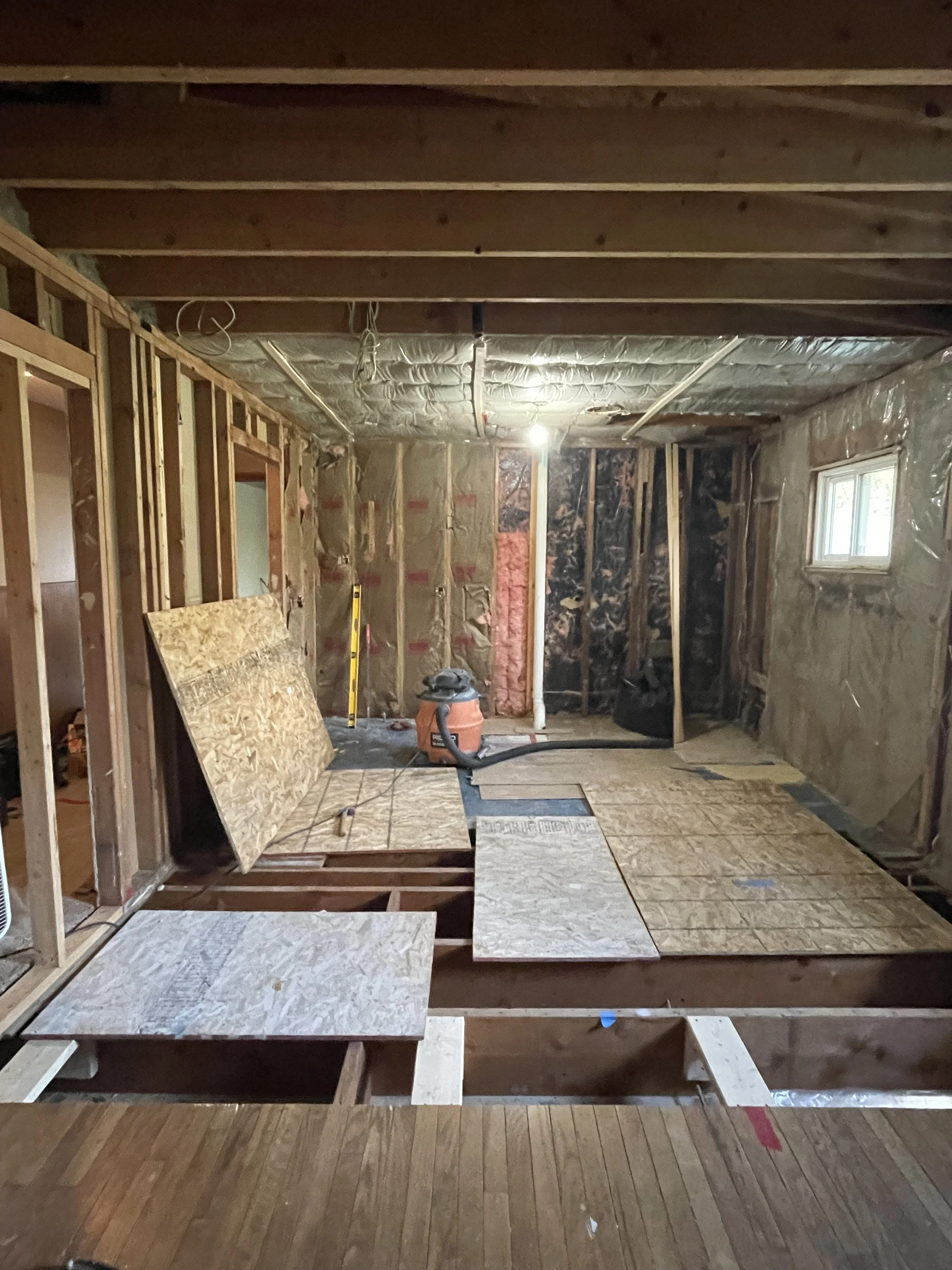 Interior of a home under renovation with exposed wall studs, insulation, and partially replaced flooring. Construction tools and materials are visible.