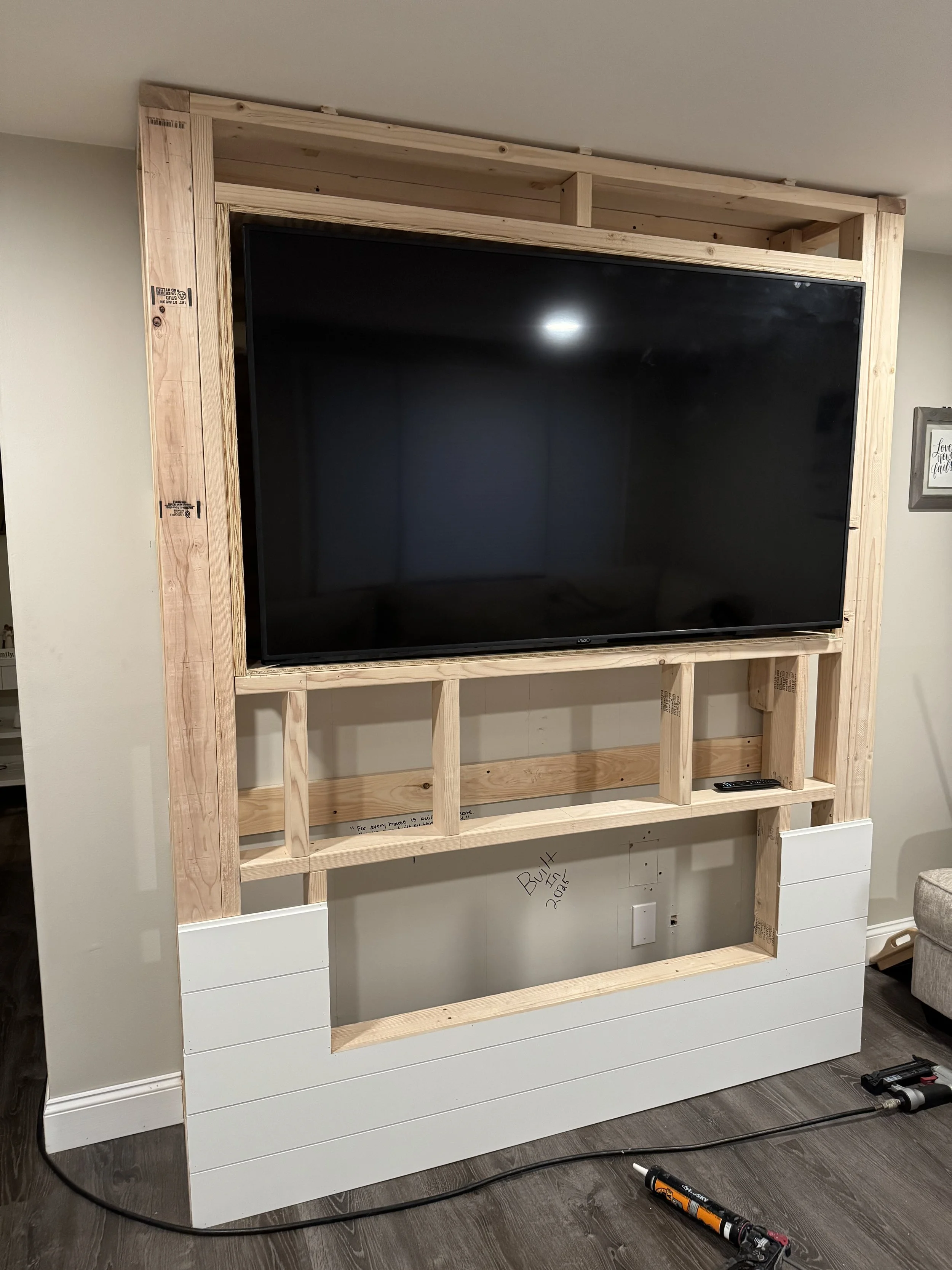 A large flat-screen TV mounted in a wooden frame under construction, with a white paneled lower section and electrical outlets beneath.