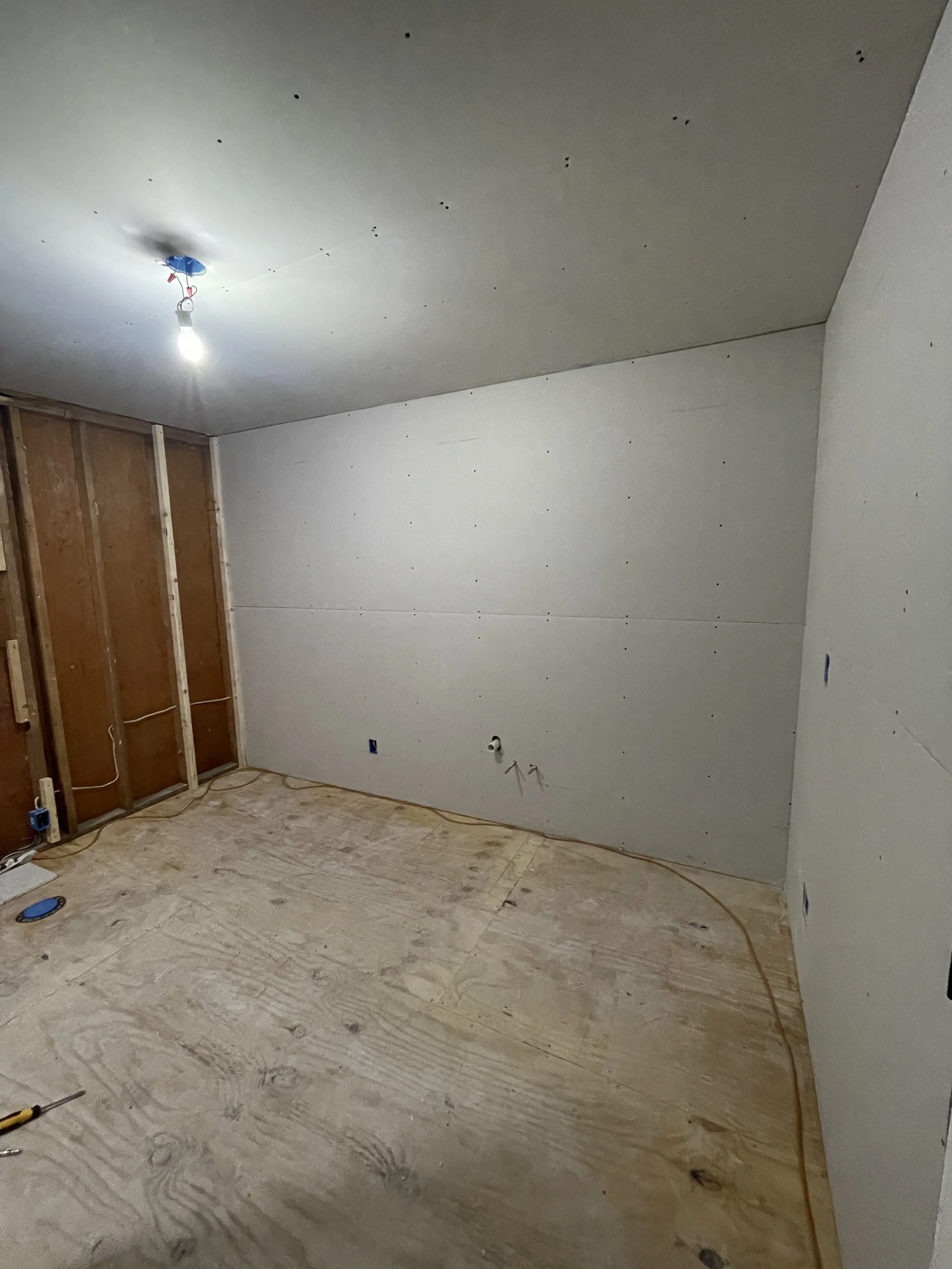 Room under construction with drywall installed on walls, exposed wooden framing, unfinished plywood flooring, and a single light bulb hanging from the ceiling.
