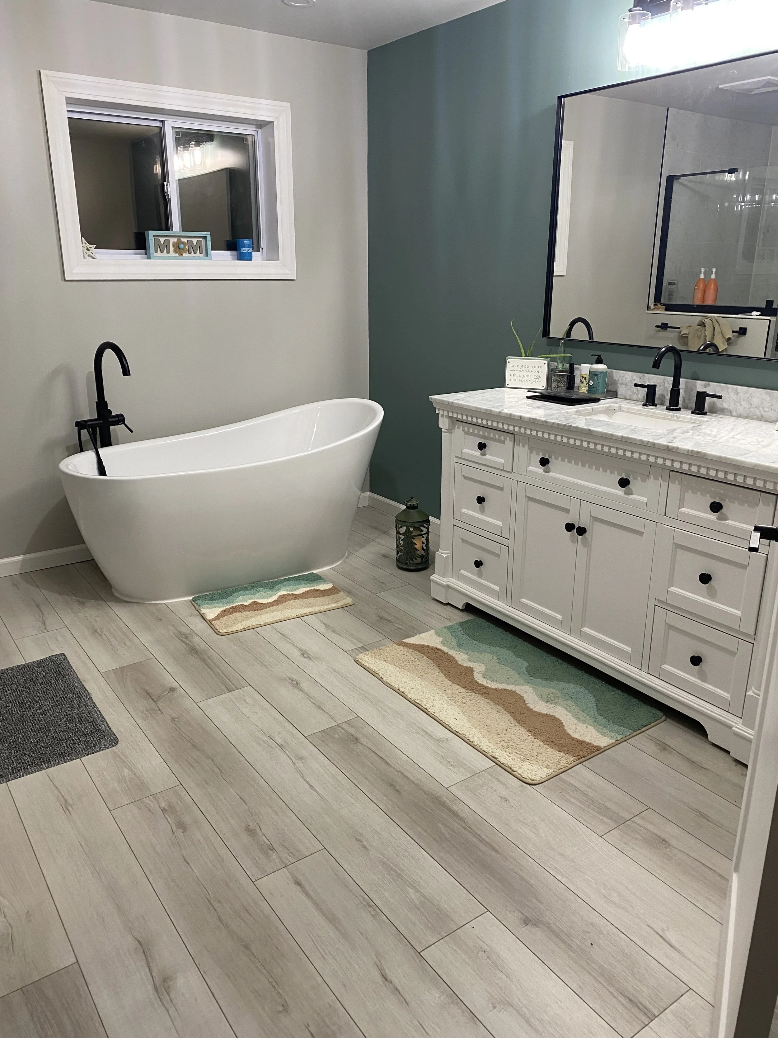 Modern bathroom with a white bathtub, a white vanity with a large mirror, and light-colored wood-like floor tiles. There are two small decorative rugs in front of the bathtub and vanity, and a lantern on the floor near the vanity. The walls are paint