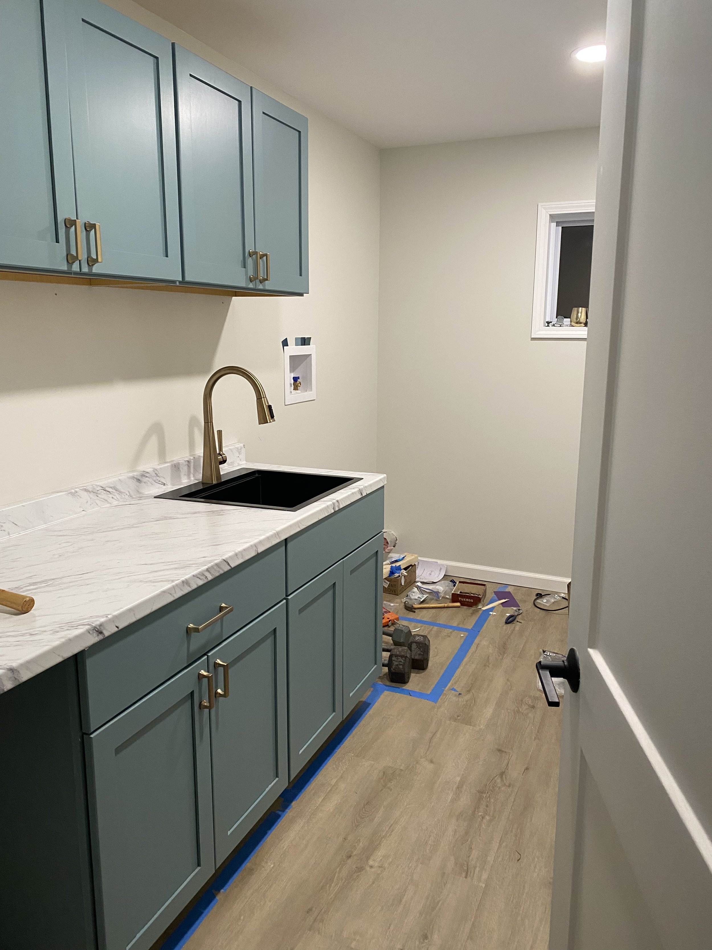 Kitchen with blue cabinets, marble countertop, black sink, and a brass faucet. There are construction tools and supplies on the floor, including a hammer, dumbbells, and boxes, indicating ongoing renovation.