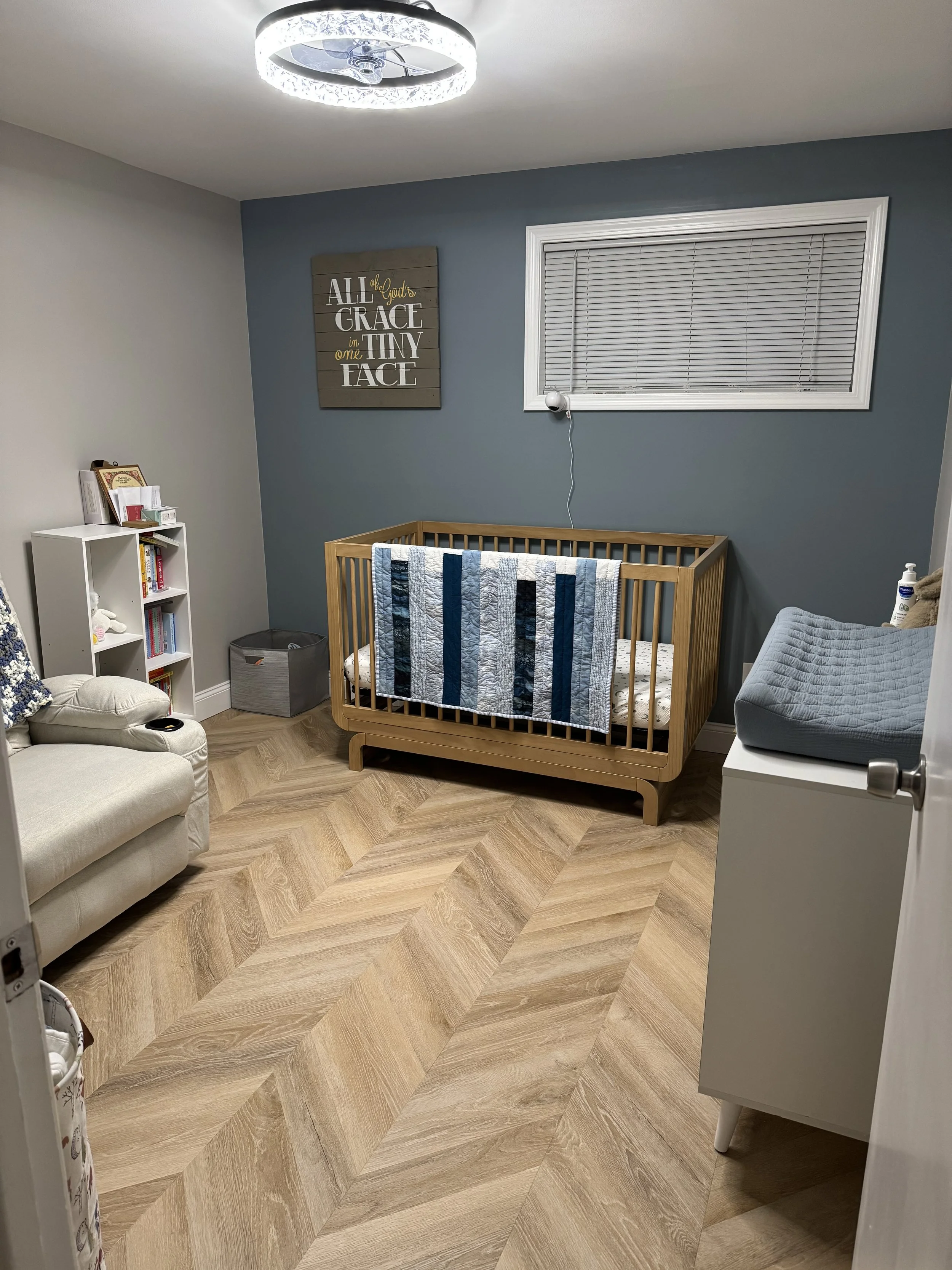 A nursery room with a wooden crib, a white armchair, a small bookcase, and a changing table with a gray changing pad. The room has wood-patterned flooring, a window with closed blinds, a wall clock, and a decorative sign on the wall that reads 'All t