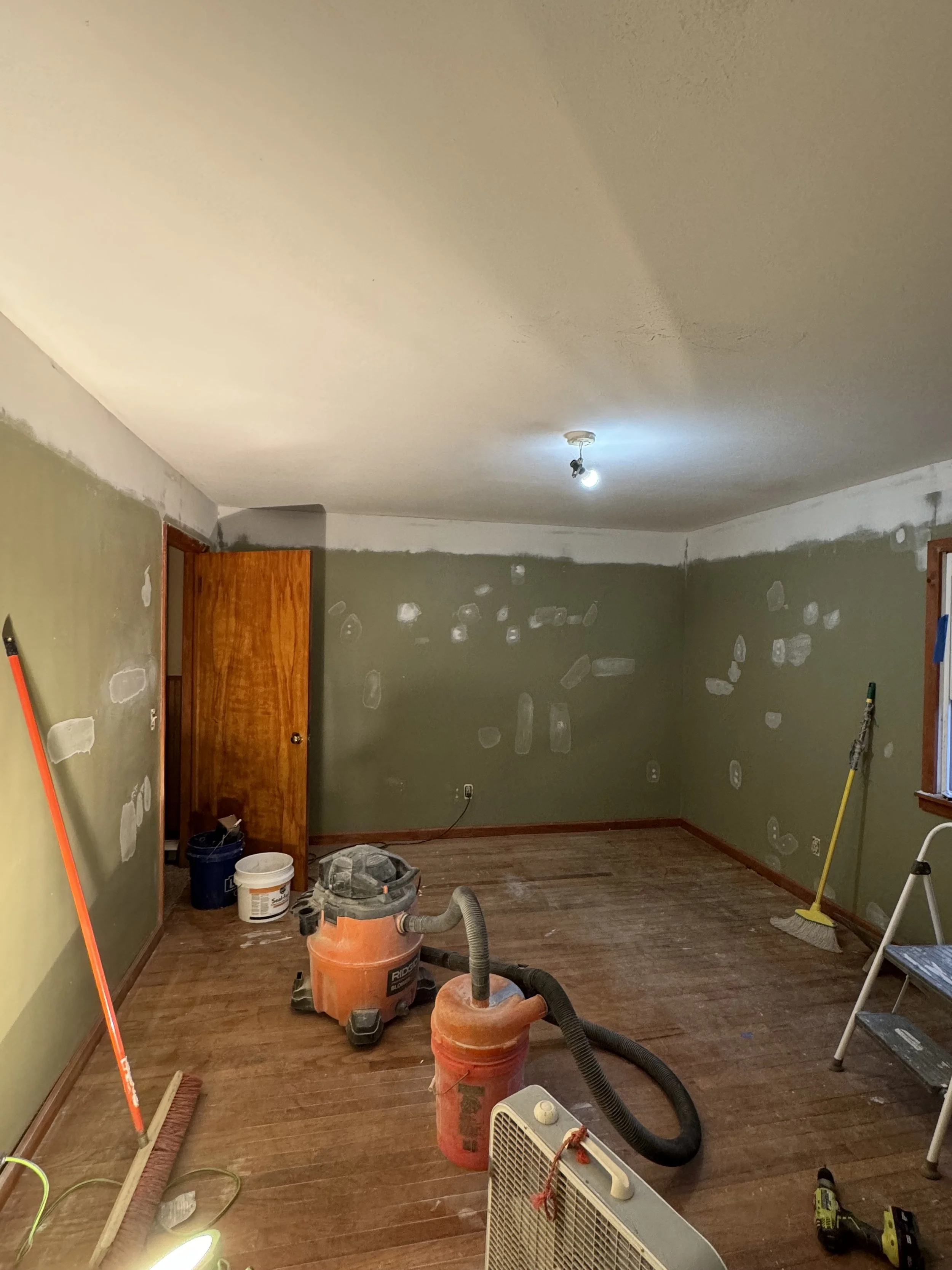 Room under renovation with walls patched for painting, vacuum cleaner, step ladder, and cleaning tools.