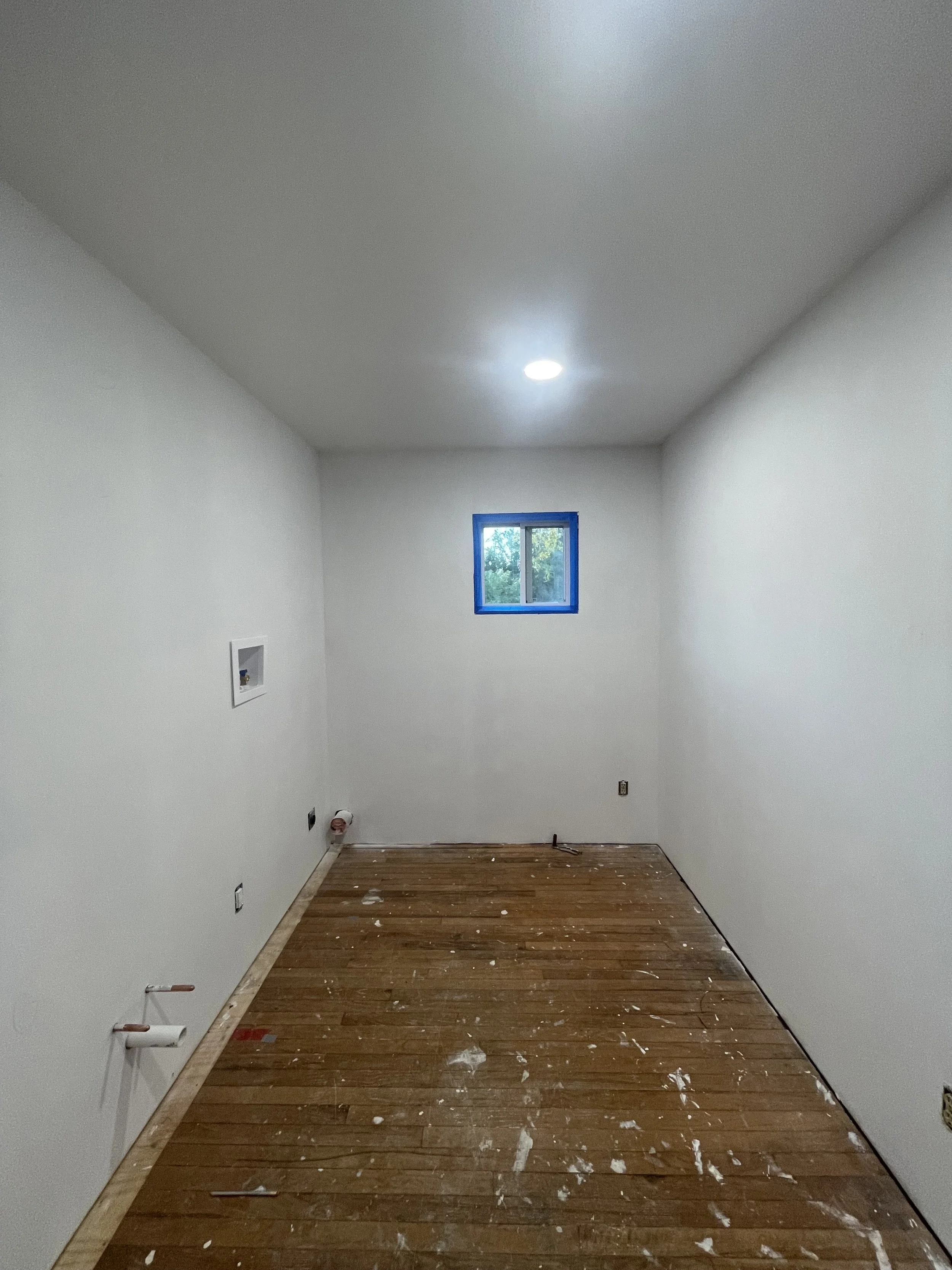 A small, empty room under construction with white walls, a small window with blue tape around it, a wooden floor with paint splatters, and some construction tools and pipes visible.