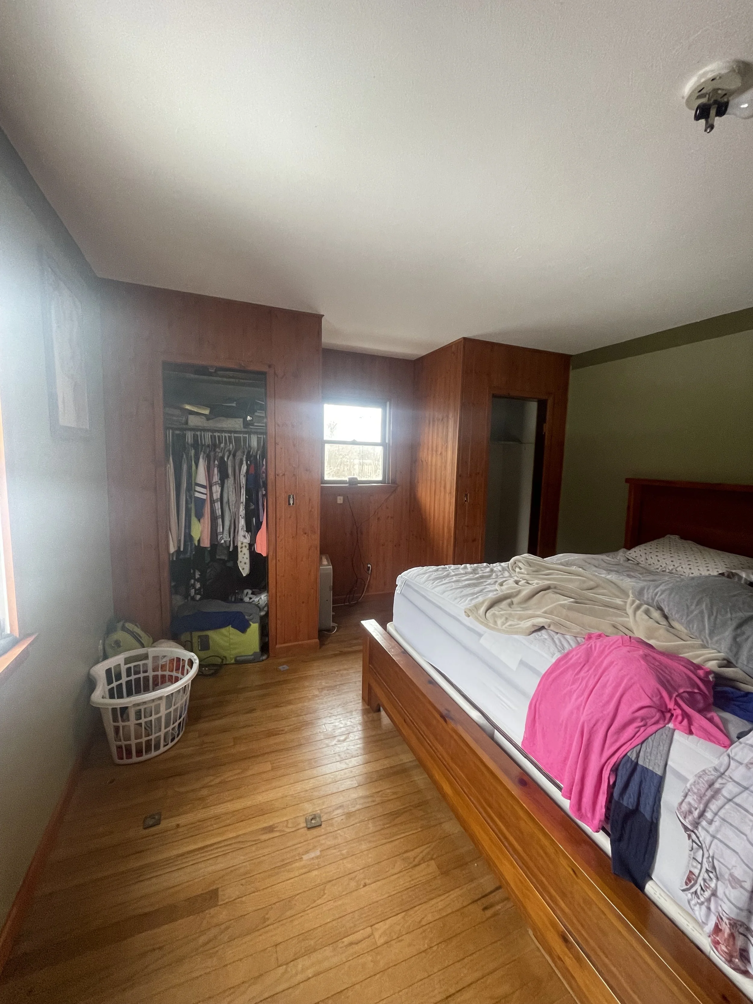 Unmade bed with various clothing items on it, open closet with clothes hanging, laundry basket on hardwood floor, small window, and wooden walls in bedroom.