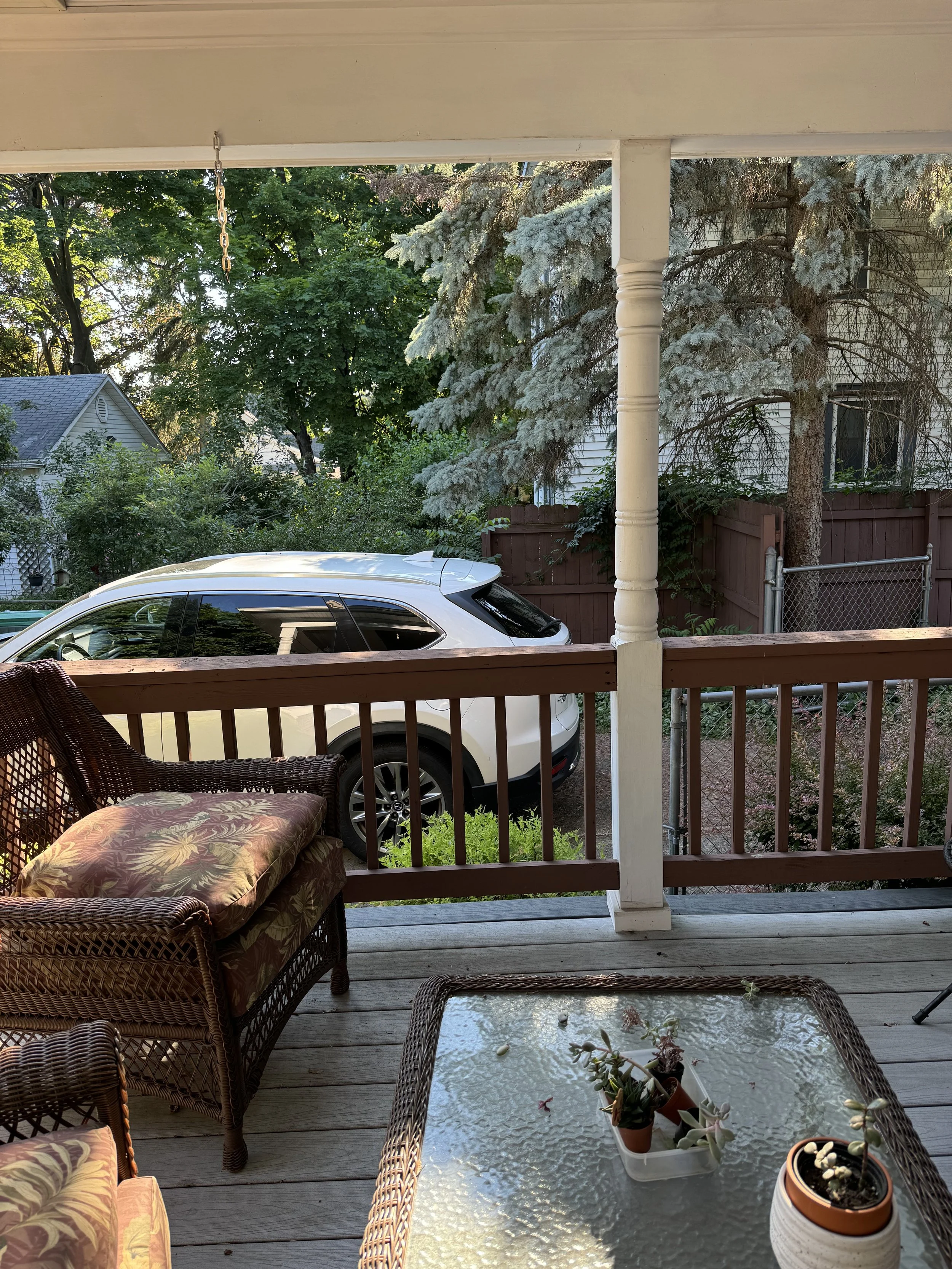 A view of a porch with wicker chairs, a glass-top table with potted plants, and a white SUV parked outside. There are trees and a wooden fence in the background.