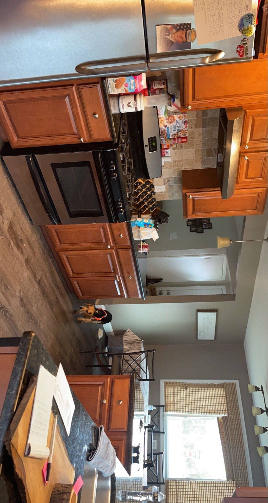 Kitchen with wooden cabinets, stainless steel refrigerator, black stove, counter with food items, a small dog sitting on the floor, and a dining area in the background with a window and window coverings.