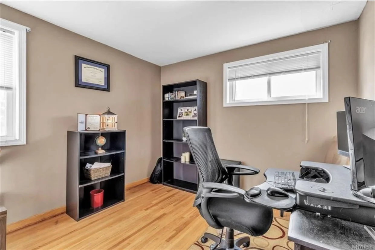 A home office with beige walls, hardwood floors, a black office chair, a desk with a monitor, keyboard, and mouse, a window with blinds, and two black bookshelves with books and decor.