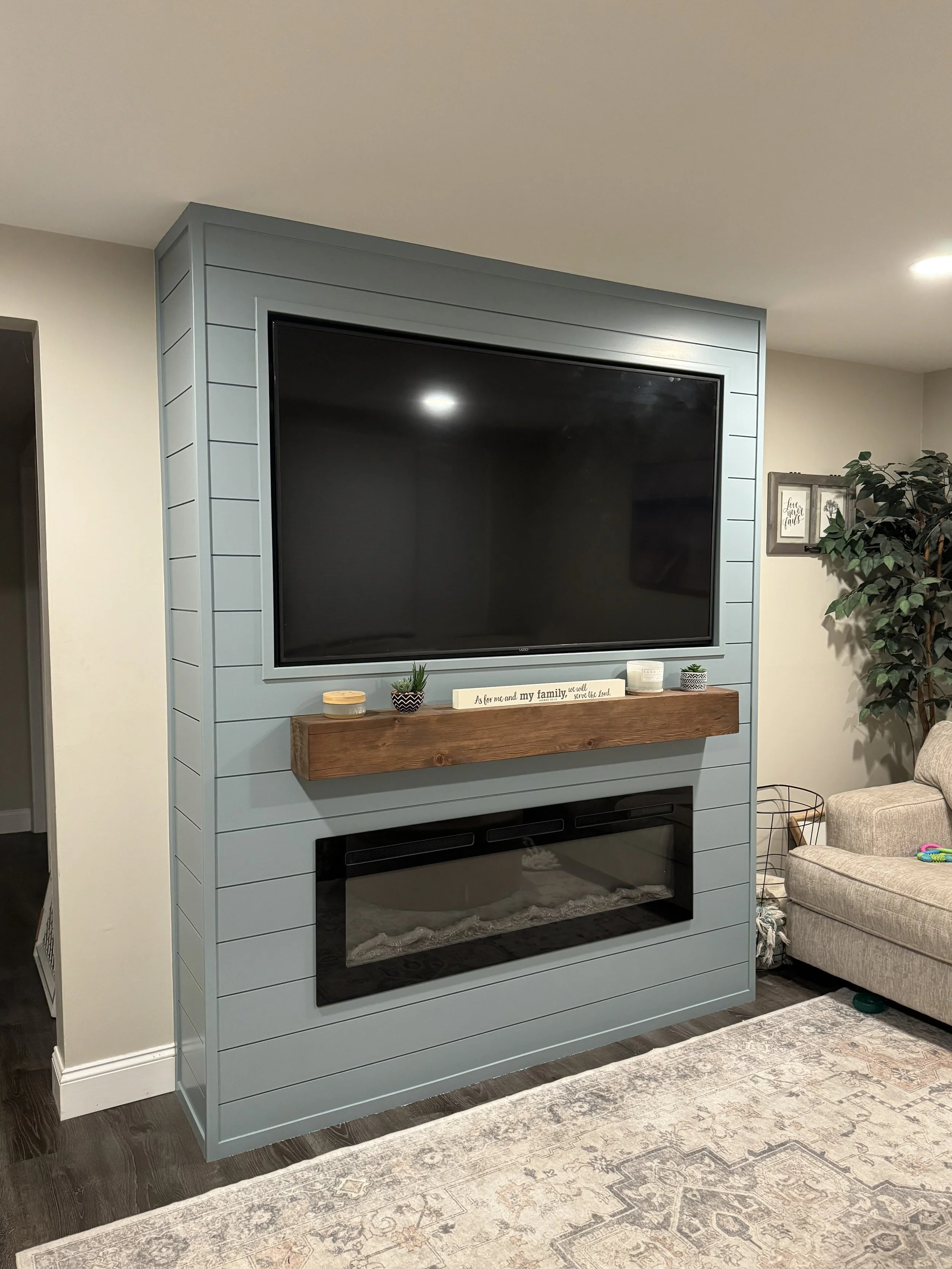 Living room corner with blue shiplap wall, mounted flat-screen TV, wooden mantel, electric fireplace, a beige sofa, and a potted plant.